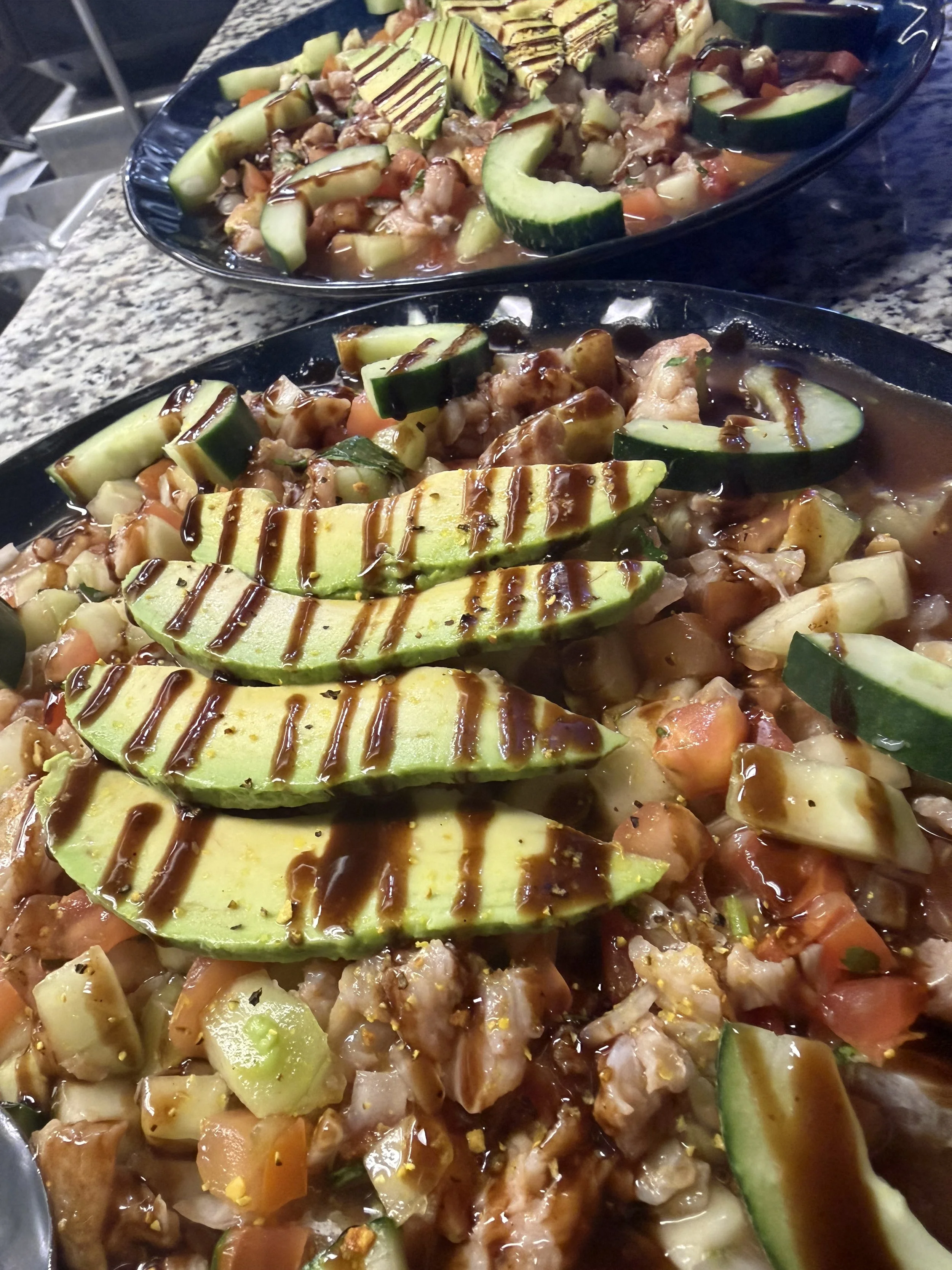 Close-up of two black serving platters of chopped vegetables including zucchini and avocado, topped with chocolate drizzle and sauce, on a granite countertop.