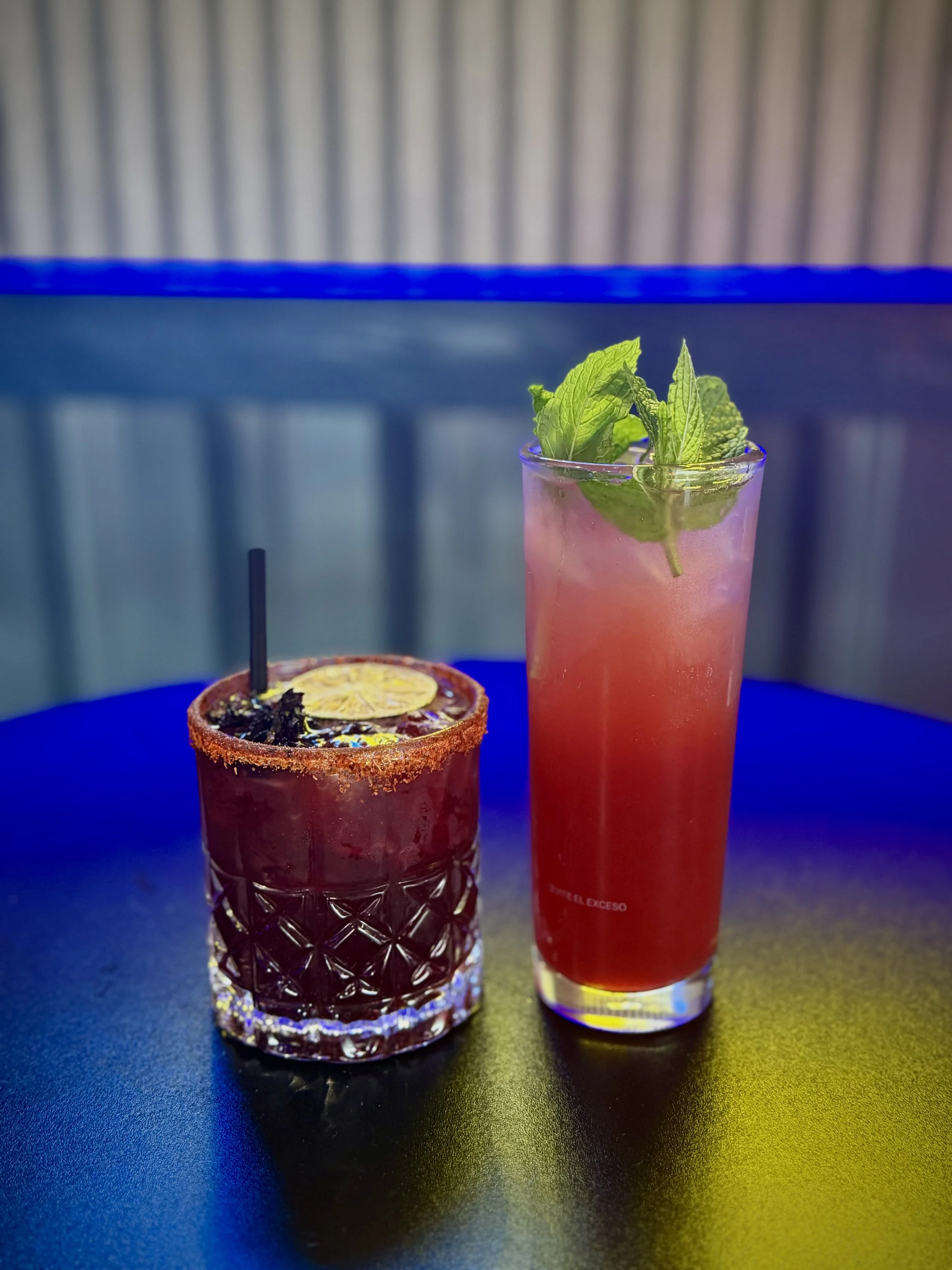 A pink beverage with mint leaves in a tall glass and a dark red drink with a lemon slice and black straw in a patterned glass.