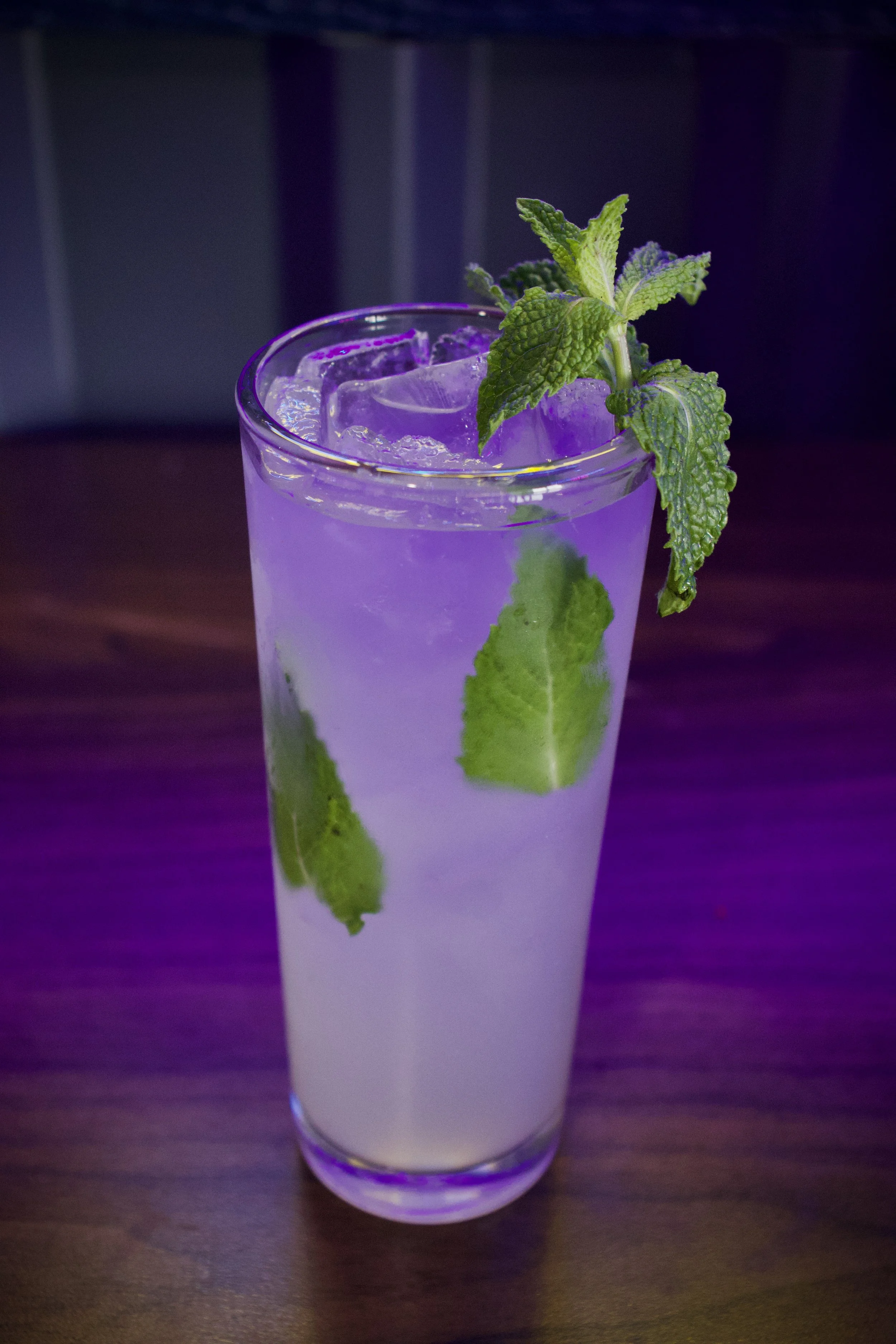 A glass of light purple cocktail with ice cubes, garnished with fresh mint leaves, on a wooden table.