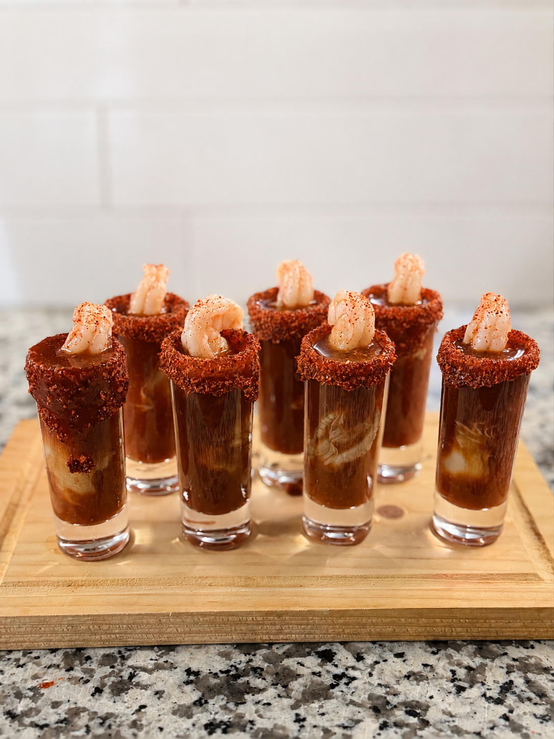 Eight shot glasses filled with a dark cocktail, garnished with a shrimp and a spicy rim, arranged on a wooden cutting board.