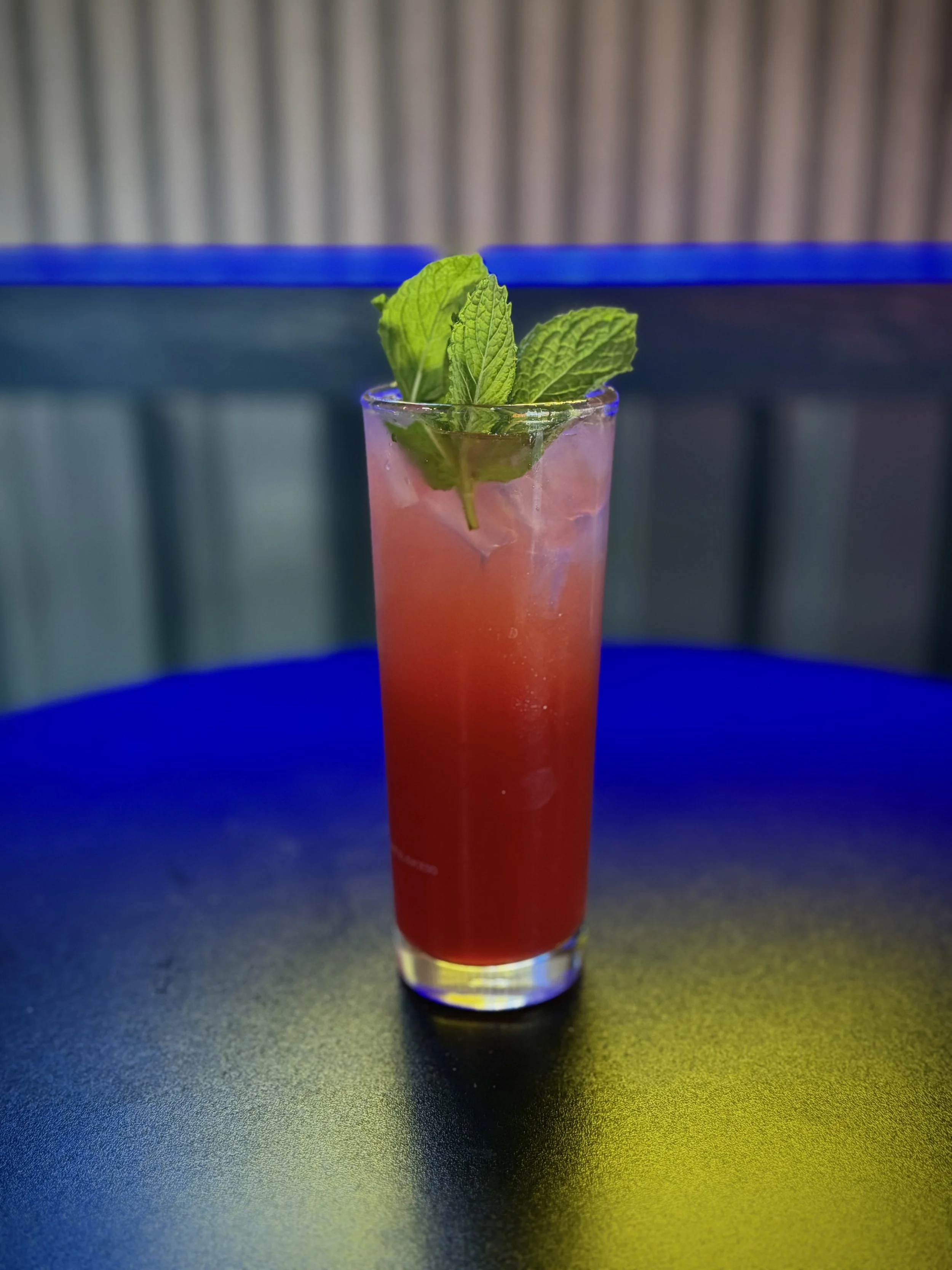 A tall glass of pink cocktail garnished with fresh mint leaves, placed on a colorful table with blue, yellow, and black sections.