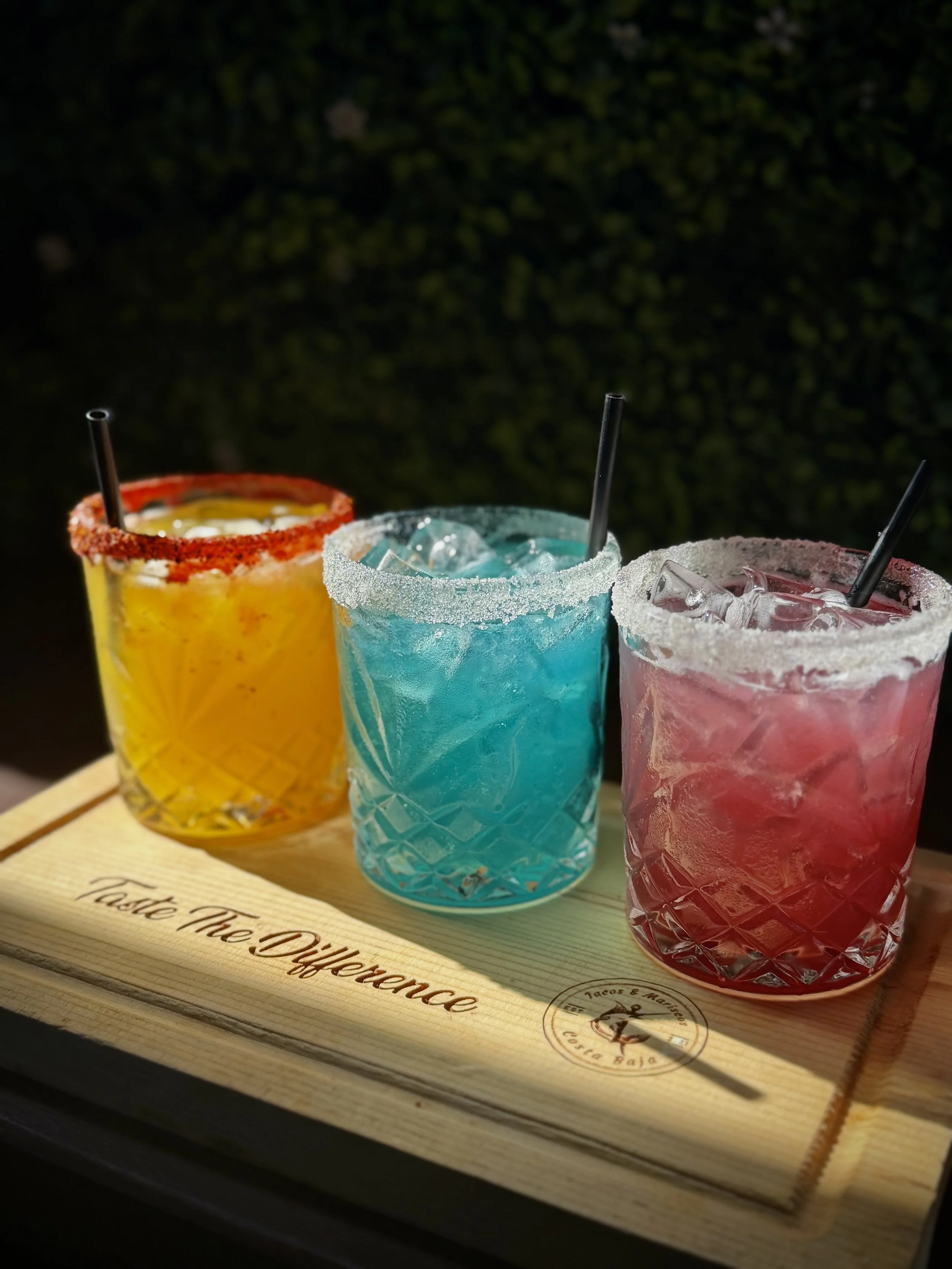 Three colorful cocktails in glasses with salted rims and straws on a wooden serving tray, against a dark background.