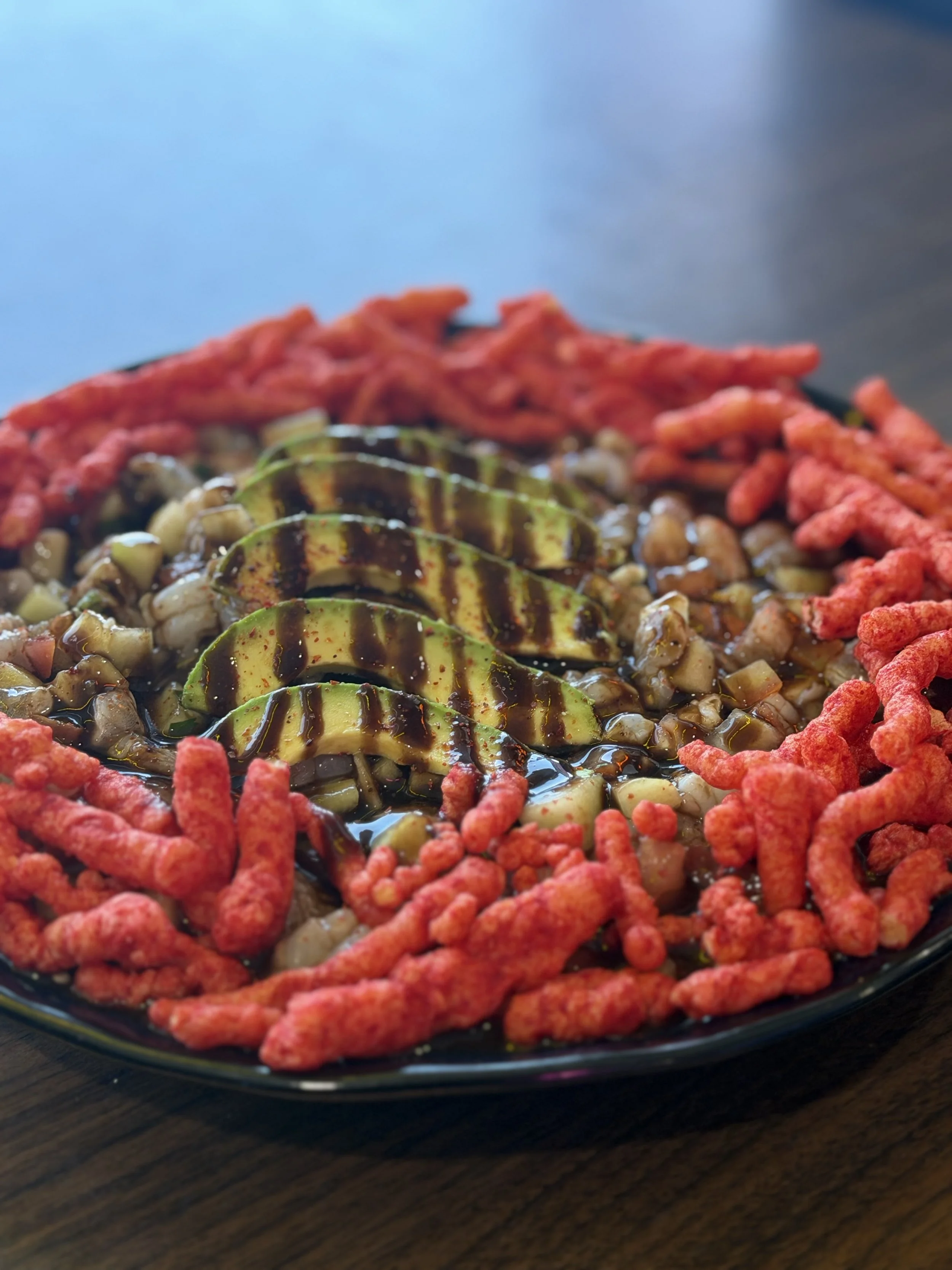 A black plate with a sushi roll topped with avocado slices drizzled with sauce, surrounded by red snack sticks and small crunchy toppings, placed on a wooden surface.
