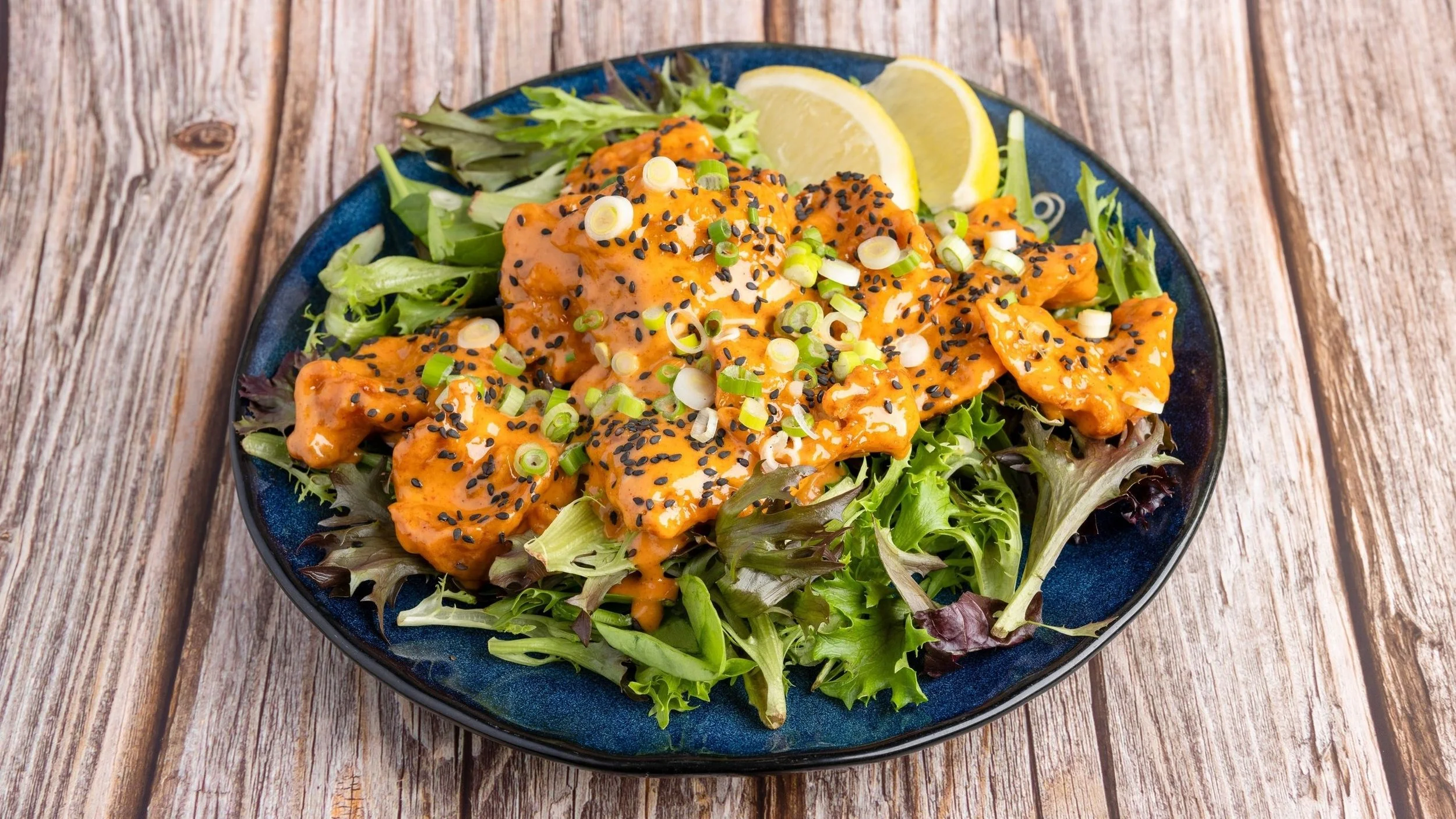 A blue plate of salad with mixed greens, topped with orange-colored chicken or seafood in a creamy sauce, garnished with chopped green onions and black sesame seeds, with lemon wedges on the side, on a wooden table.