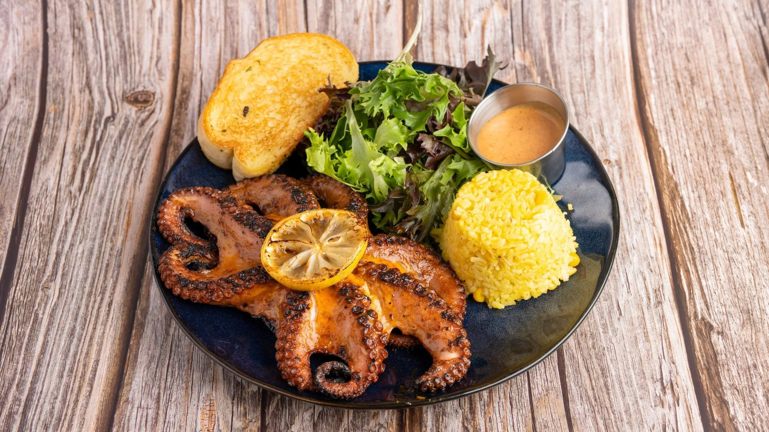 Plate of grilled octopus with lemon, yellow rice, salad, garlic bread, and dipping sauce on a wooden table.