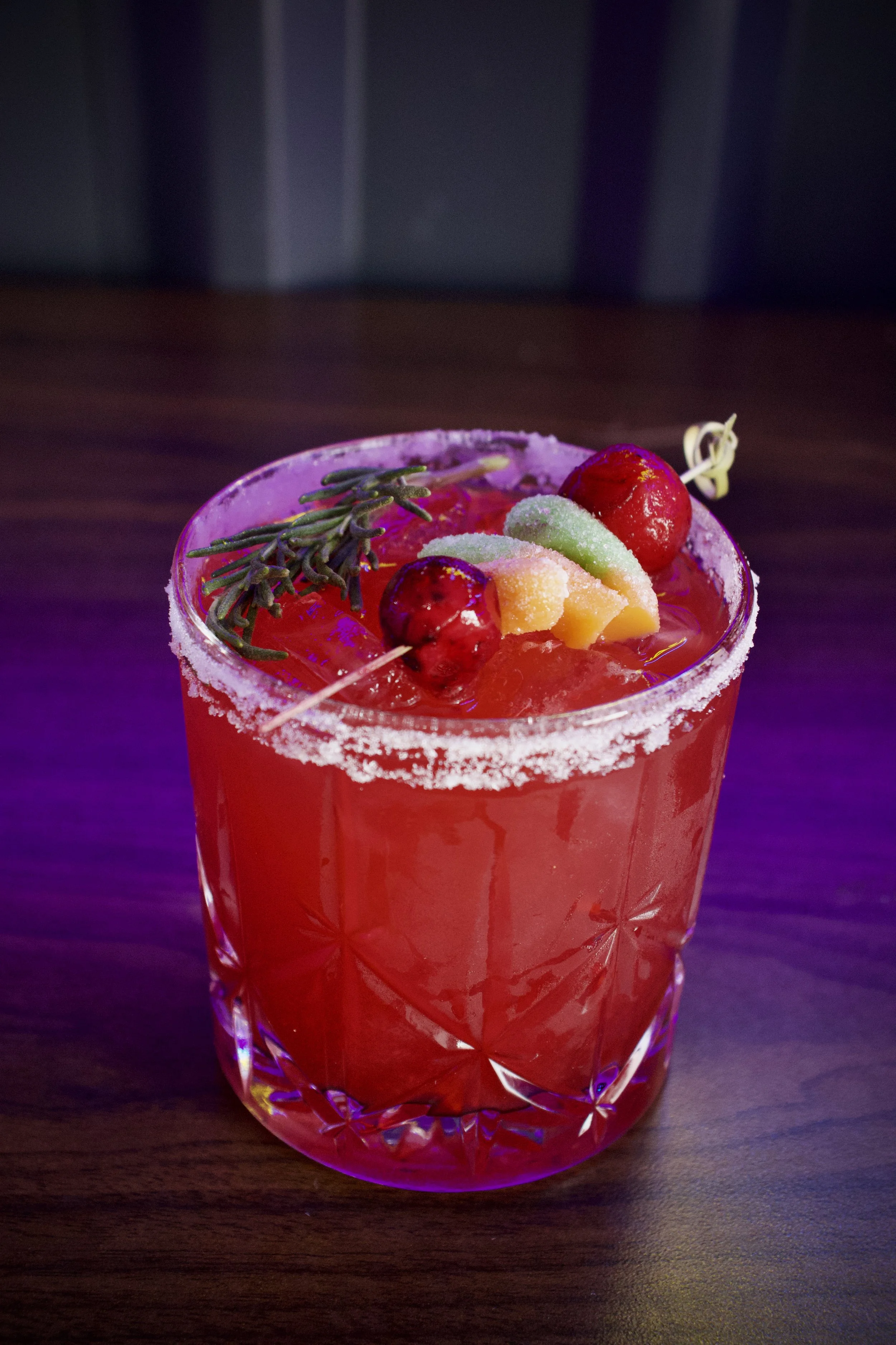 Close-up of a red cocktail in a decorative glass with a salted rim, garnished with a sprig of rosemary, two cherries, and a slice of lime on top. The drink is set on a dark wooden surface.