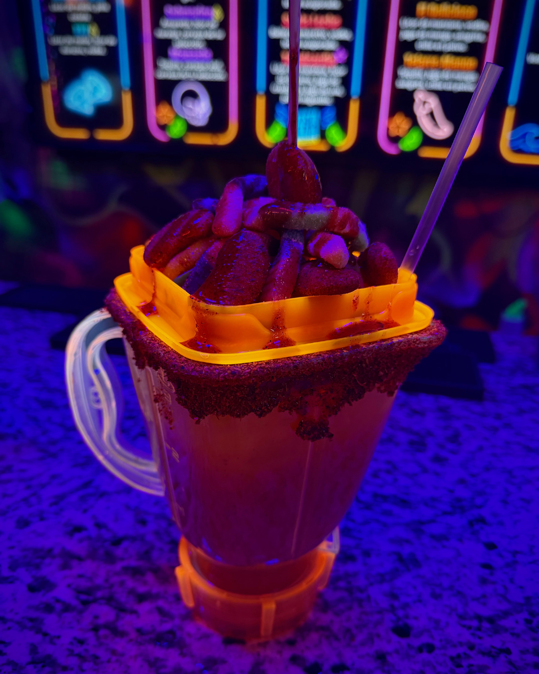 A neon-lit candy and snack display featuring a large cup filled with various candies and snacks, with colorful illuminated signs in the background.