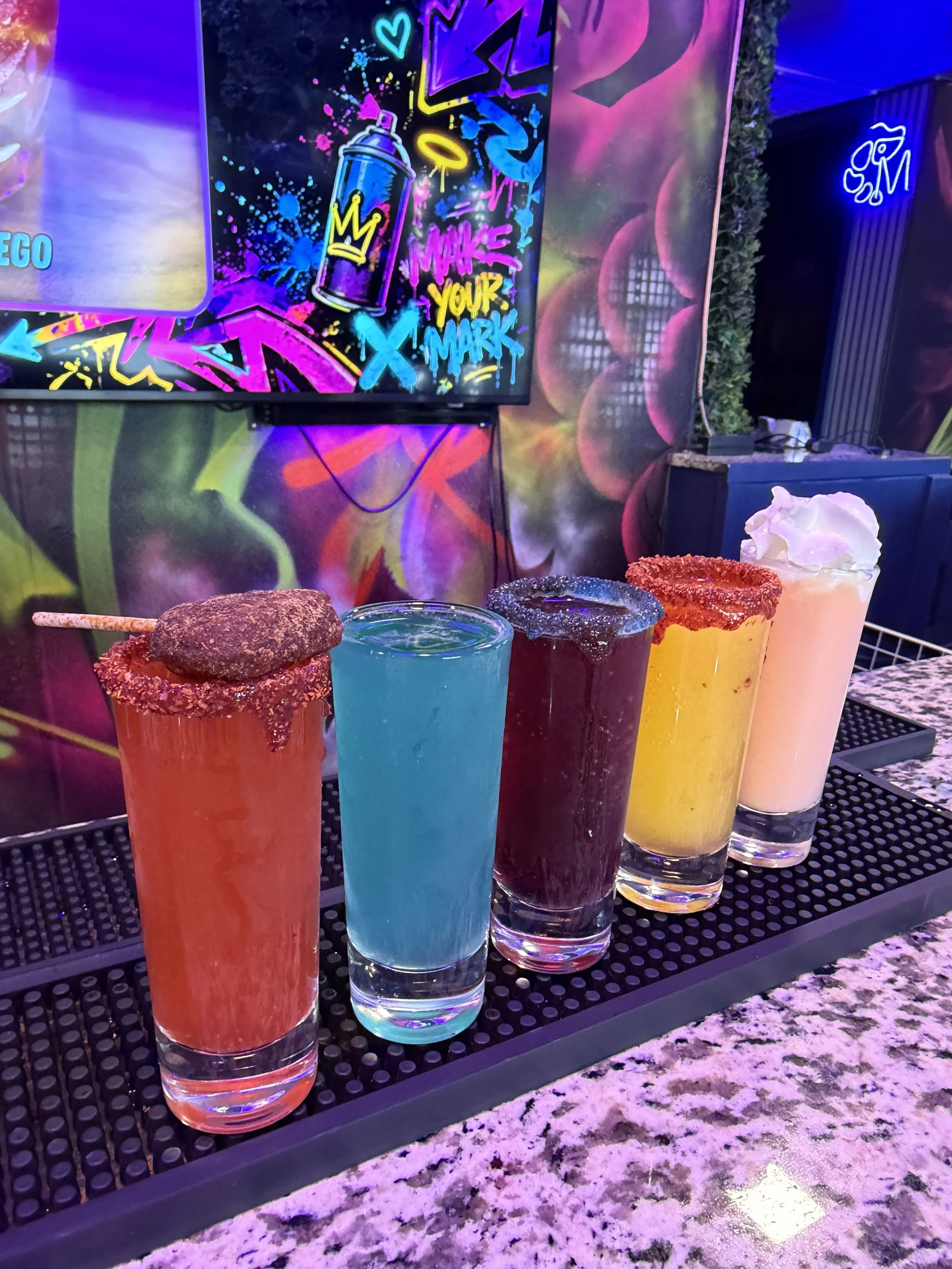A row of colorful cocktails topped with various garnishes on a black tray, with vibrant graffiti-style neon signs and artwork in the background.