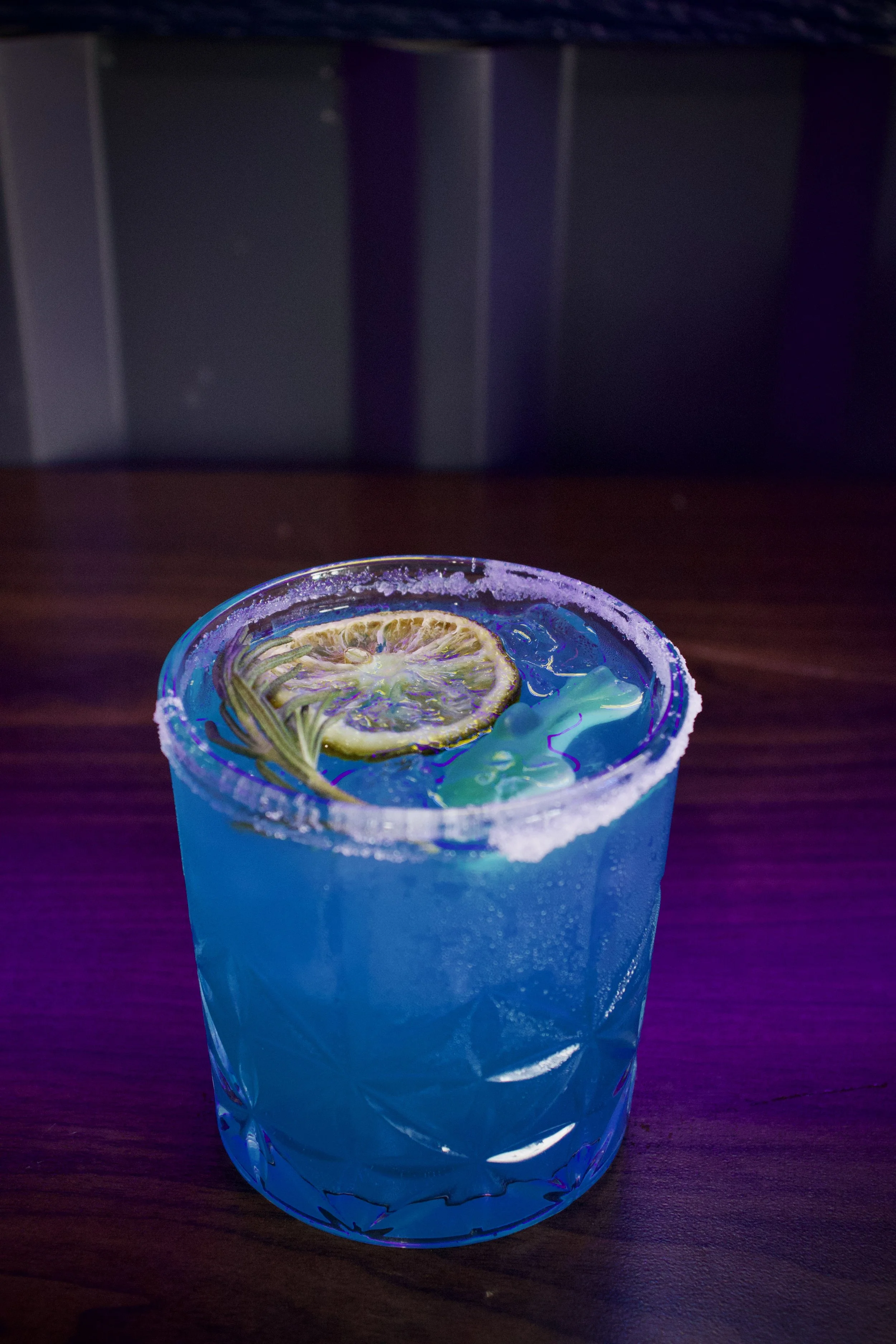 A blue cocktail glass with a salted rim, containing a brightly colored drink garnished with a dried lemon slice.