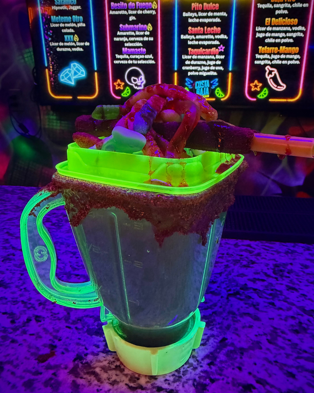 A neon-lit photo of a large transparent cup with a green lid, filled with a dark liquid, topped with a green plastic tray containing various fried snacks and a skewer. The background features a colorful menu on a neon sign with drinks and their ingre