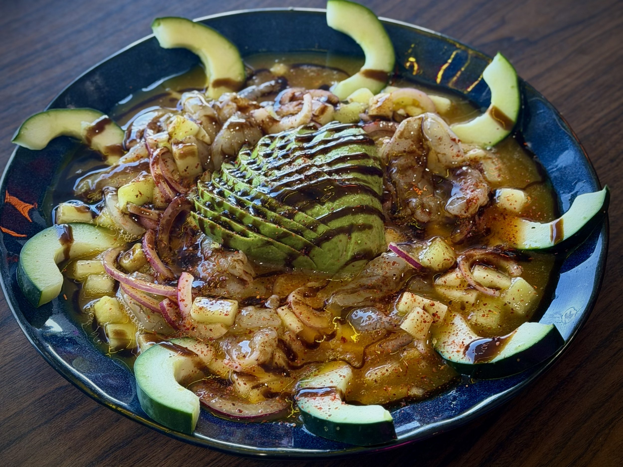 A black bowl with slices of cucumber arranged around the edge, filled with a soup or stew with sliced onions, avocado slices, and a drizzle of dark sauce on top.