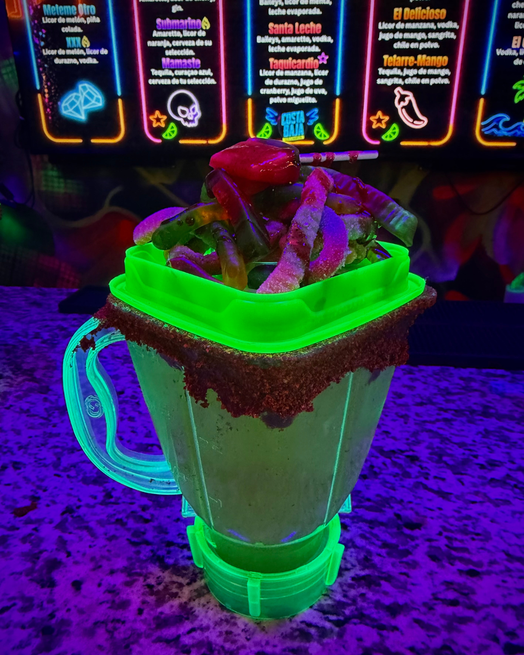 Glow-in-the-dark beverage with gummy worms and a red gummy on top, served in a neon-green holder with a glittery border, set on a brightly lit counter with neon menu signs in the background.