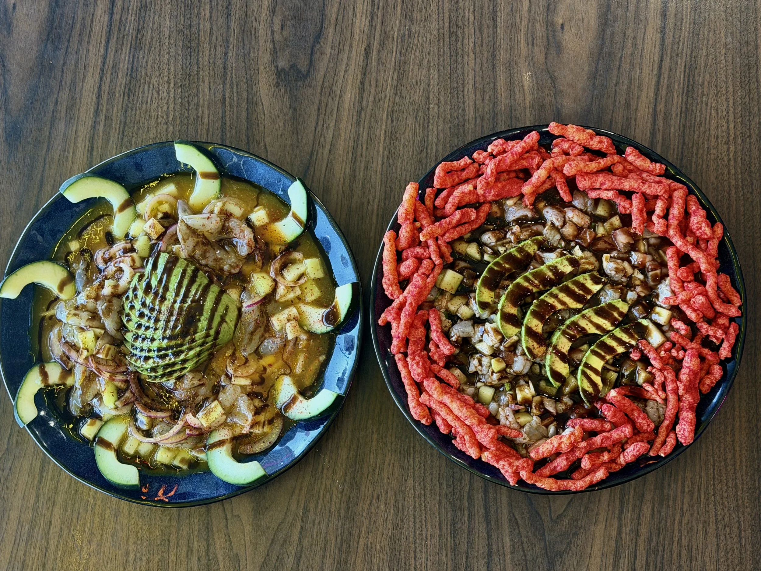 Two bowls of Asian cuisine on a wooden table. The left bowl contains sliced cucumbers, onions, and a brown sauce. The right bowl contains sliced cucumbers, diced vegetables, and red snack sticks around the edge.