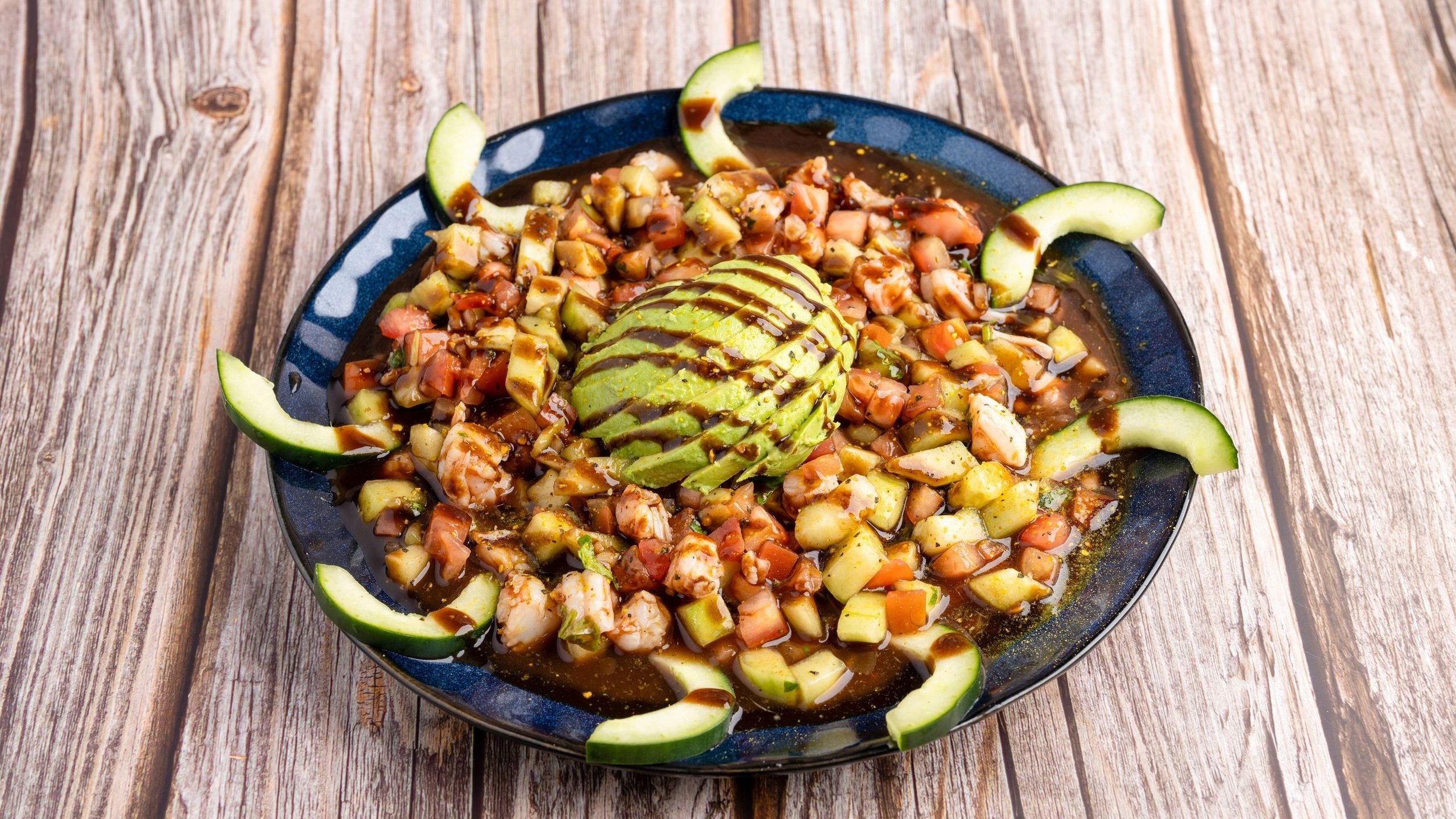 A plate of ceviche garnished with sliced avocado and cucumber on a wooden table.