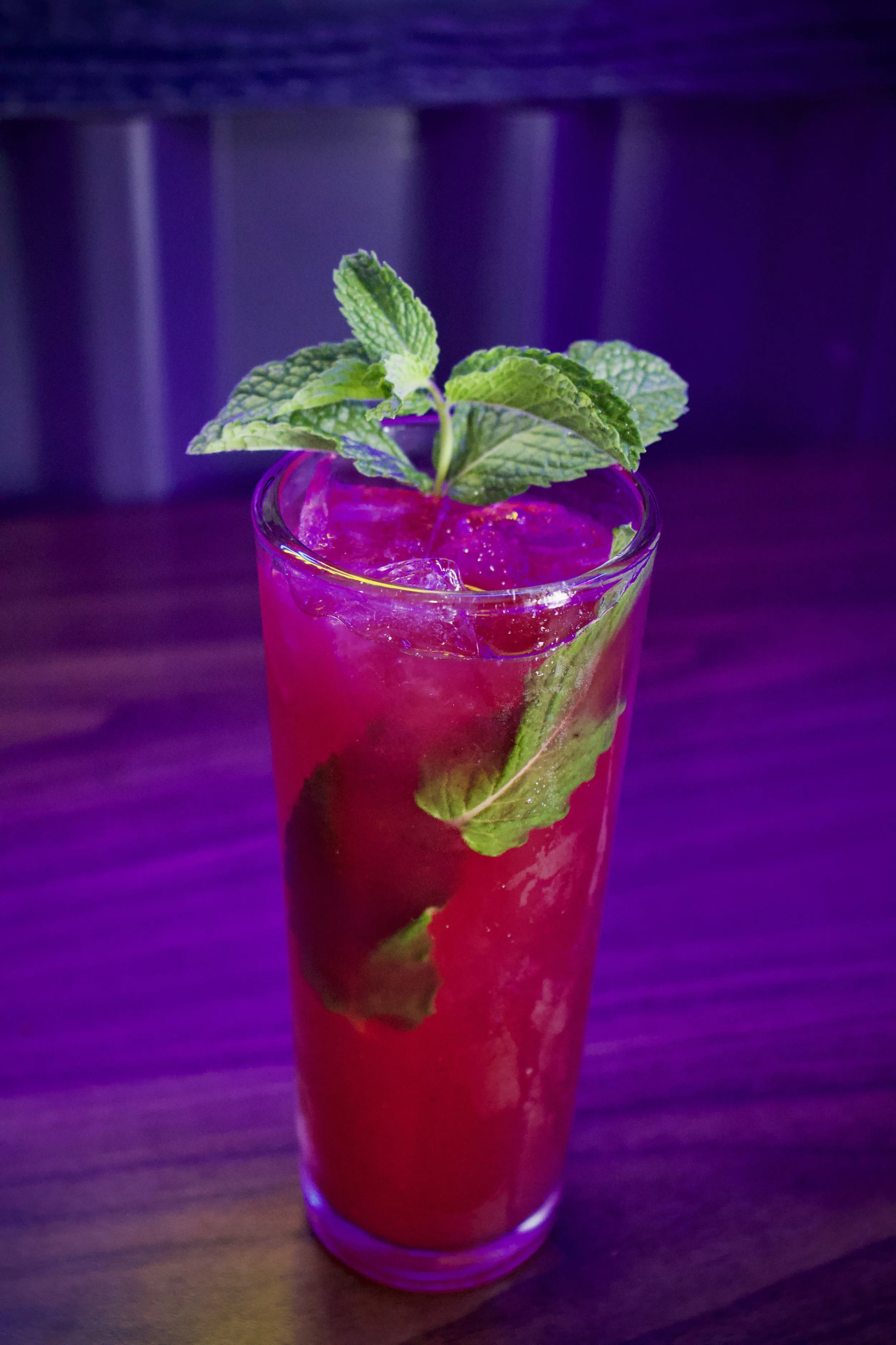 A colorful cocktail in a tall glass with ice cubes, garnished with fresh mint leaves and a lime slice, set on a wooden surface with dim purple and blue lighting.