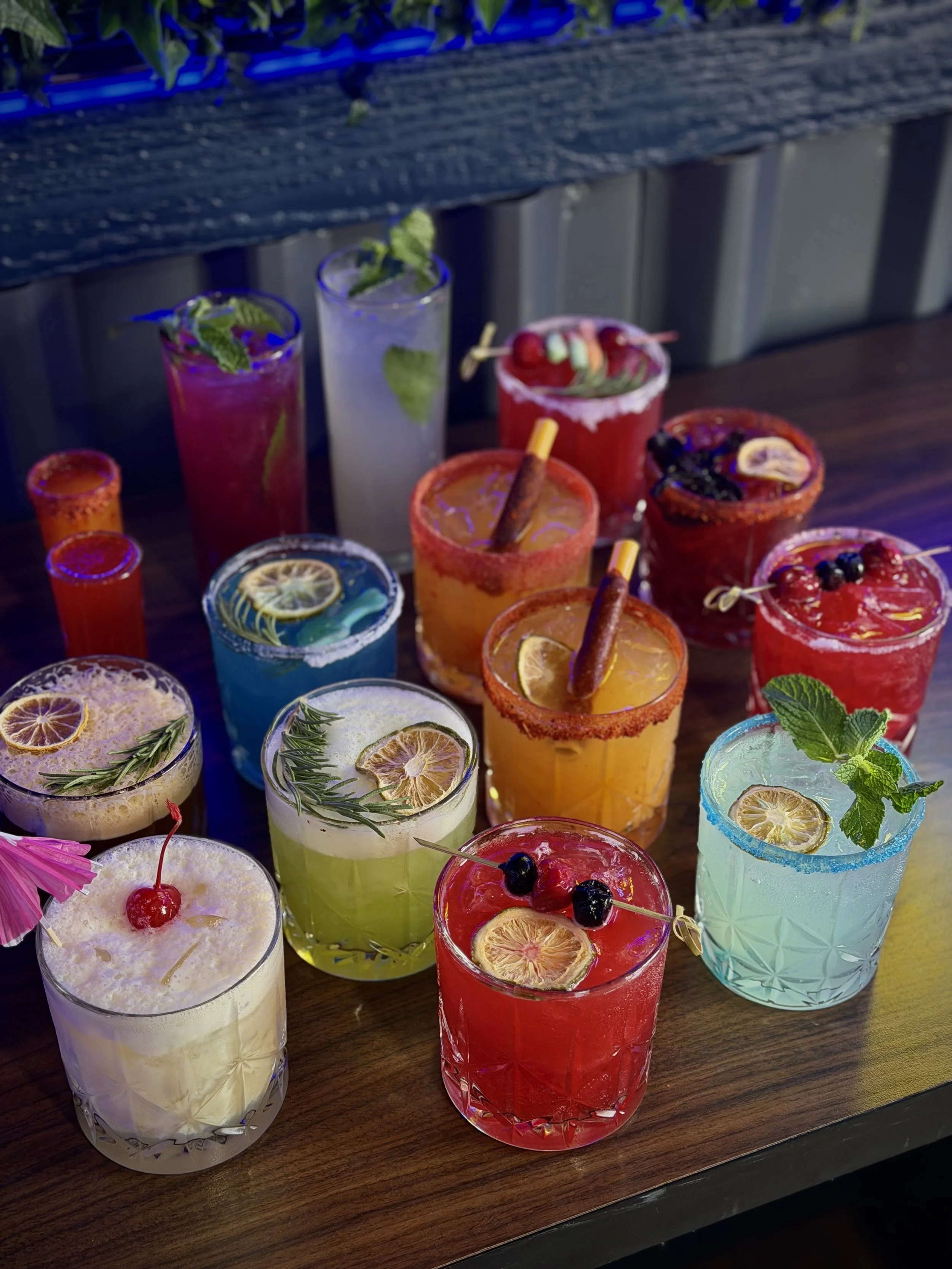 Colorful cocktails with lemon and cherry garnishes on a dark wooden table at a bar or restaurant.