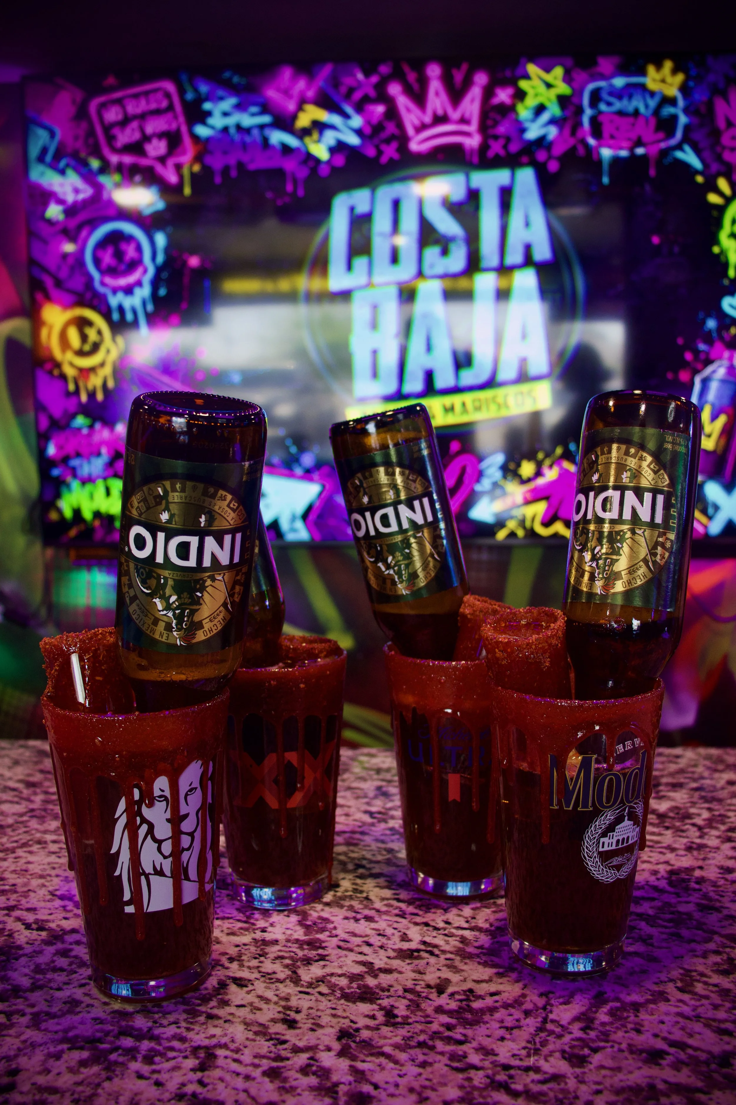 Three glasses of dark drink topped with small bottles with black caps labeled "Indio" sit on a speckled pink countertop. In the background, a colorful neon sign displays the words "Costa Haja" and various graffiti-style designs and text.
