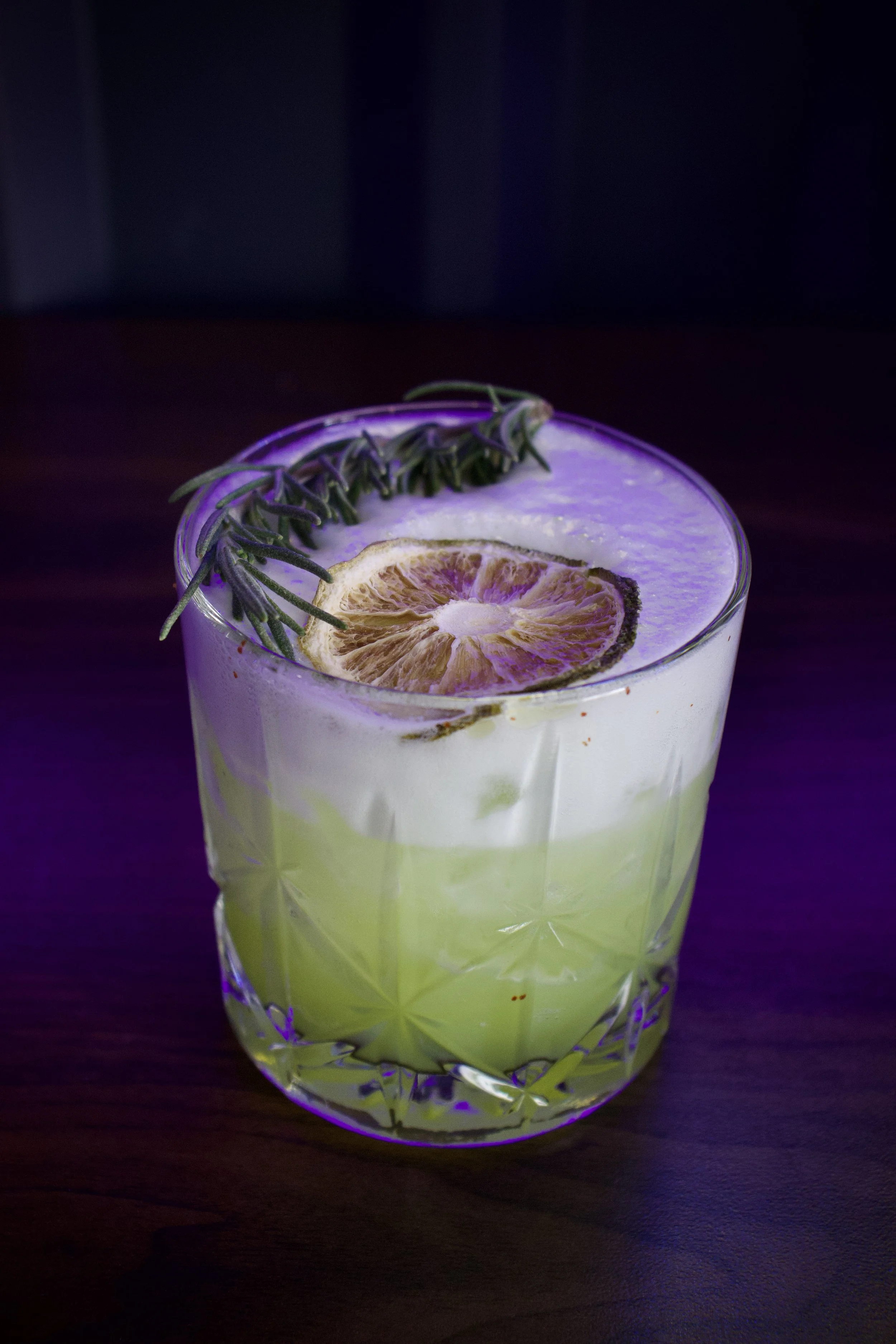 A glass with a pale yellow-green drink garnished with a dried citrus slice and a sprig of rosemary, set on a dark surface.