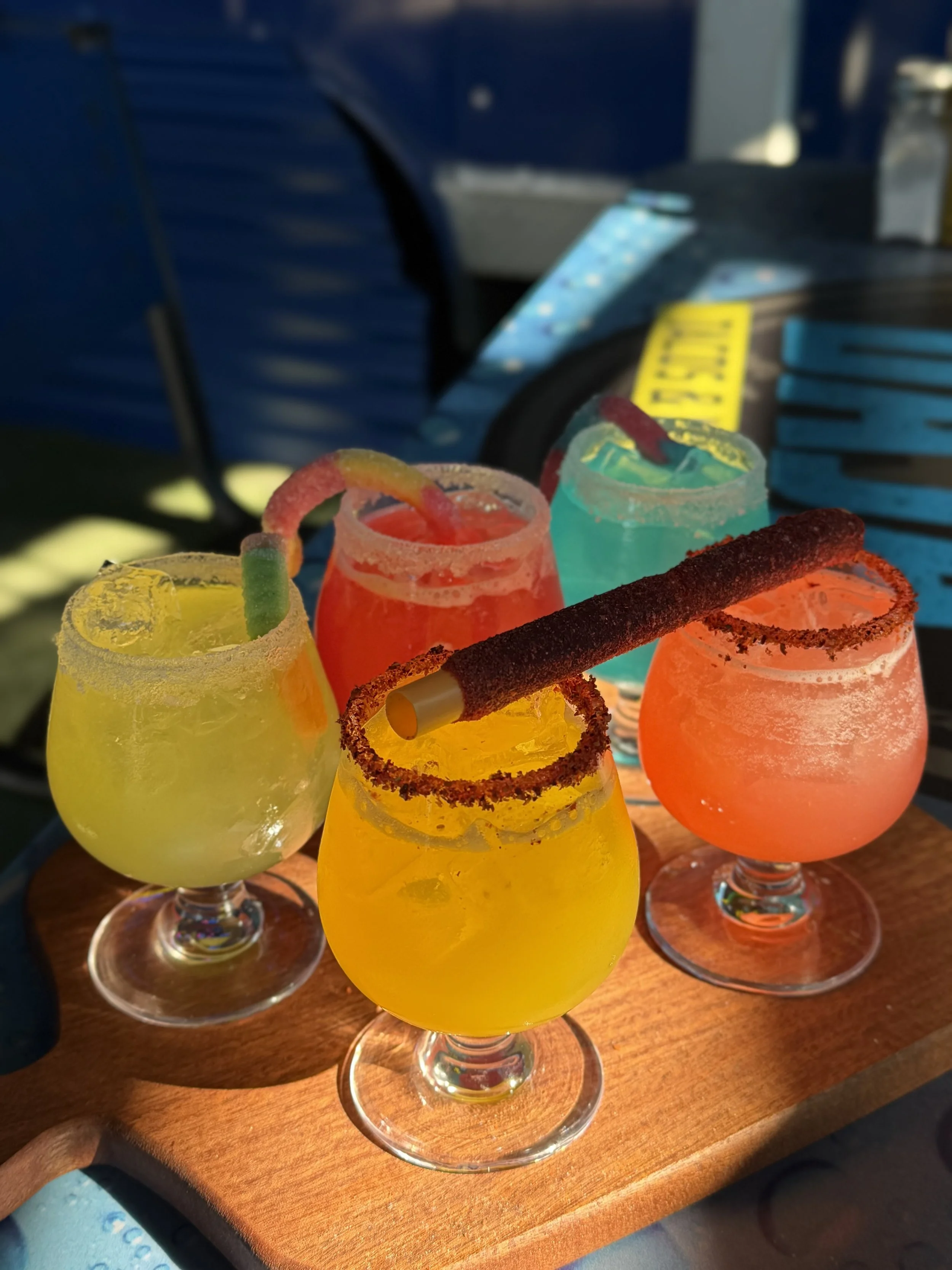 Four colorful cocktails with salt and sugar rimmed glasses and garnishes, served on a wooden tray.