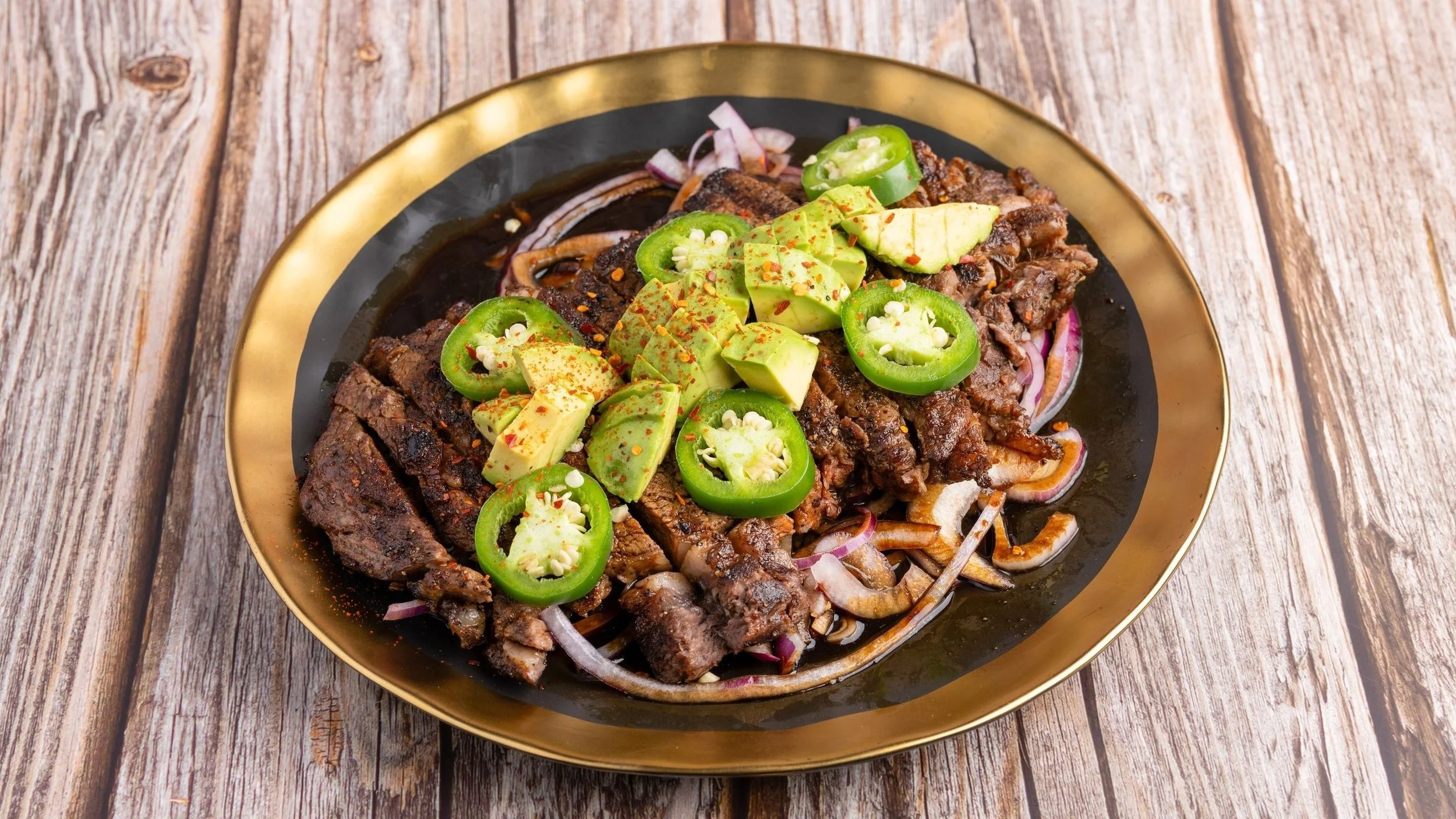 Plate of grilled steak topped with sliced avocados, jalapeño peppers, and seasoned with chili flakes, served on a bed of sliced onions on a black and gold plate, placed on a rustic wooden table.