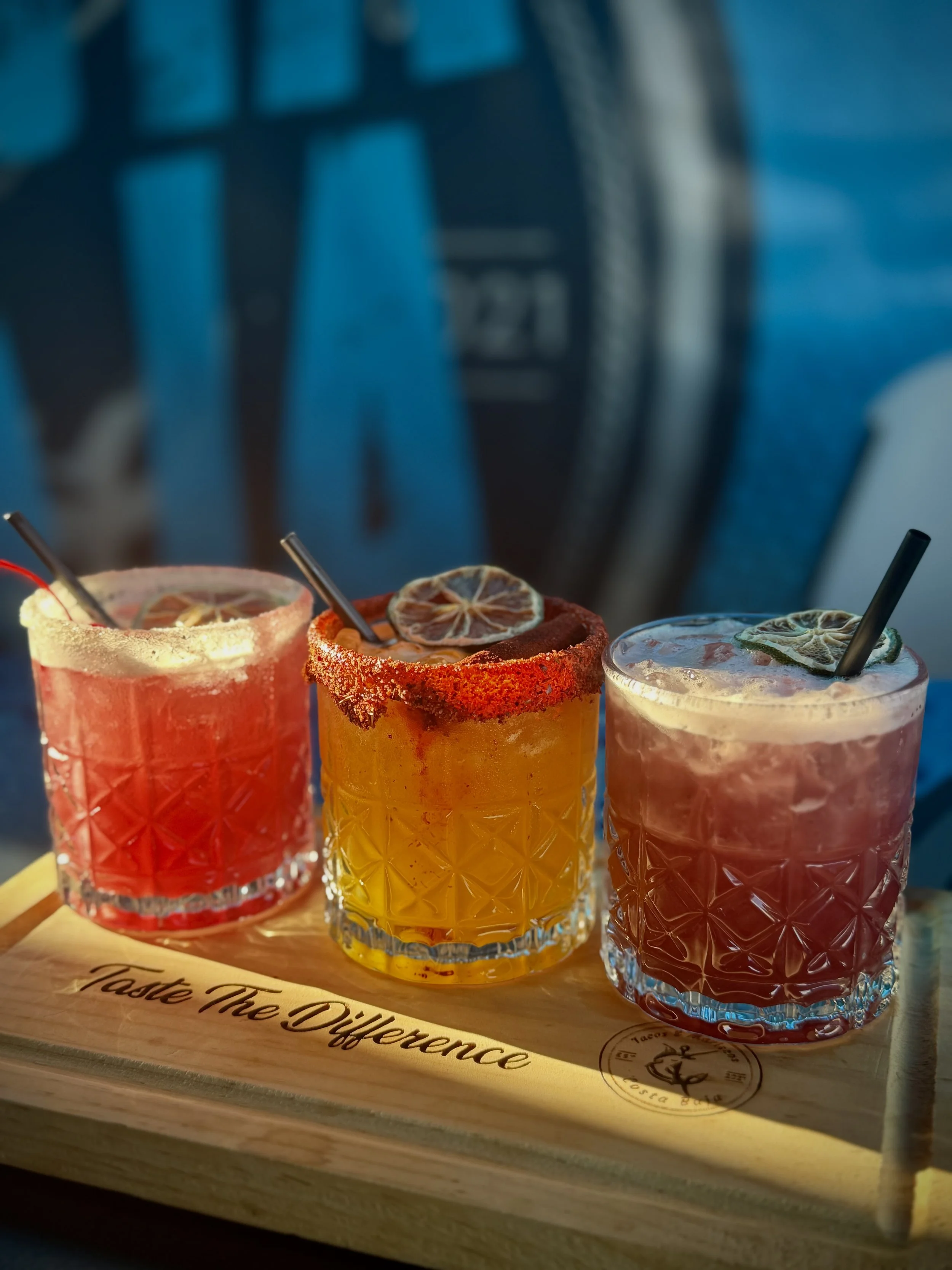 Three colorful cocktails with dried citrus garnishes and straws on a wooden serving board with the phrase 'Taste The Difference' engraved, against a blue background.