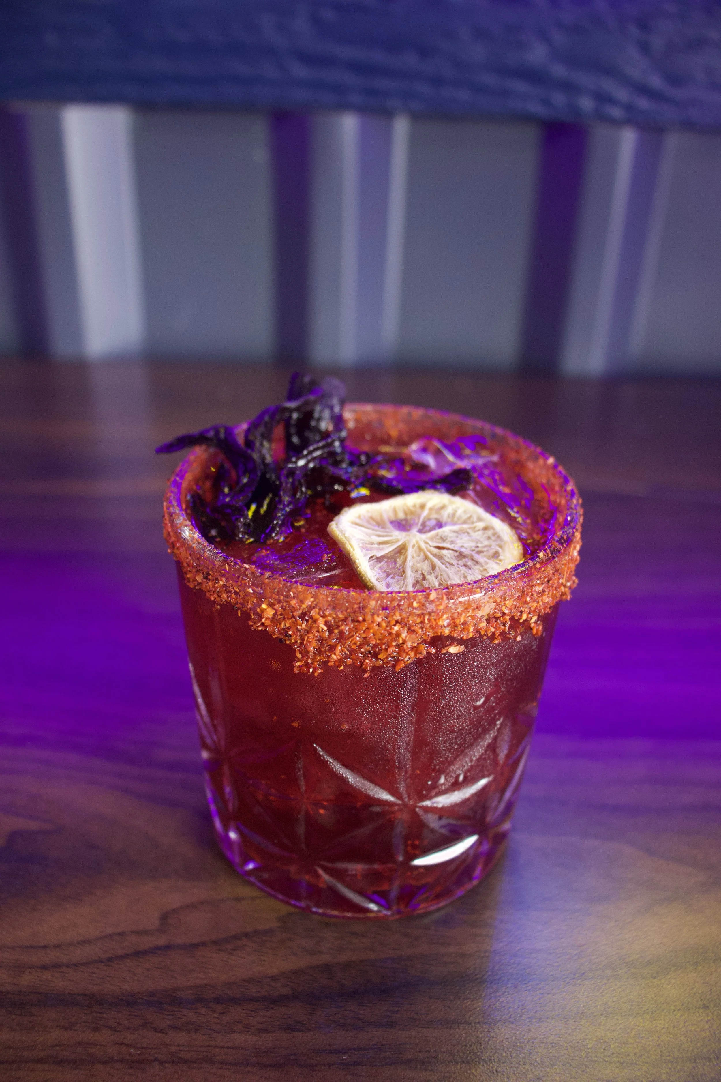 A close-up of a red cocktail in a glass with a salted rim, garnished with a dried lemon slice and purple herbs, sitting on a wooden surface with purple lighting.