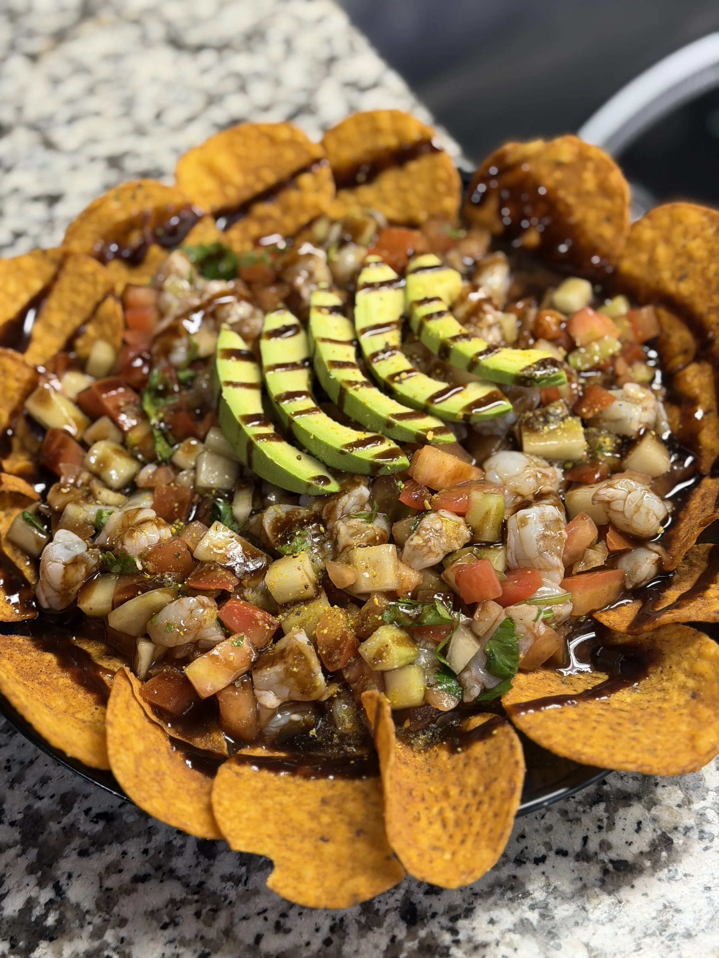 Close-up of a plate of loaded nachos with tortilla chips, topped with diced tomatoes, lettuce, avocado slices, shredded chicken, and drizzled with dark sauce or dressing, set on a granite countertop.