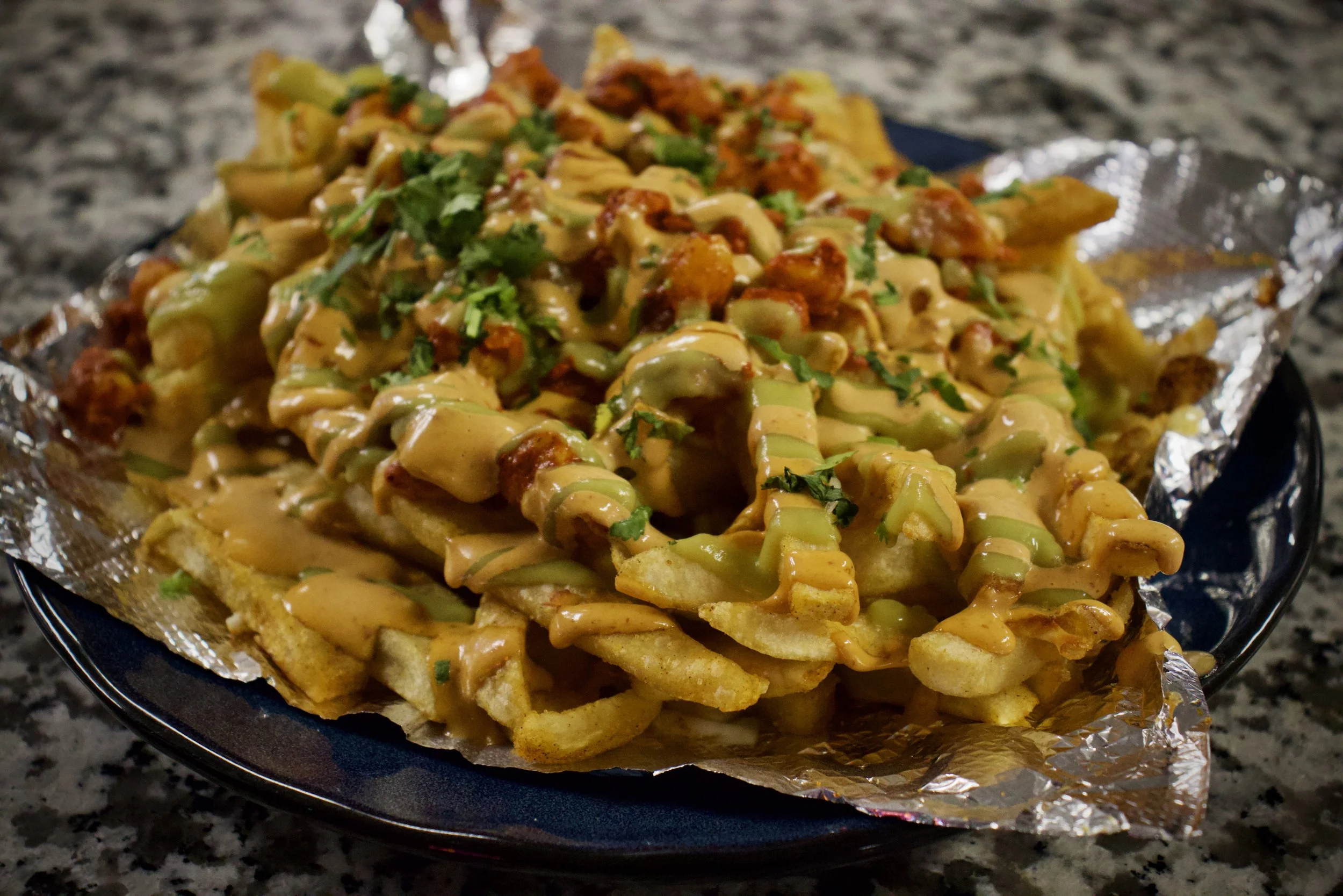 Plate of loaded French fries topped with cheese, spicy meat sauce, fresh chopped herbs, and drizzled with sauce.
