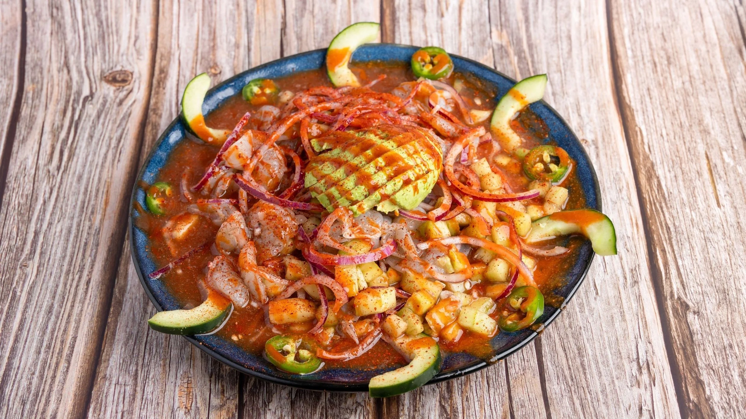 A bowl of shrimp ceviche garnished with avocado slices, cucumber, jalapeño slices, red onion, and a spicy red sauce on a wooden surface.