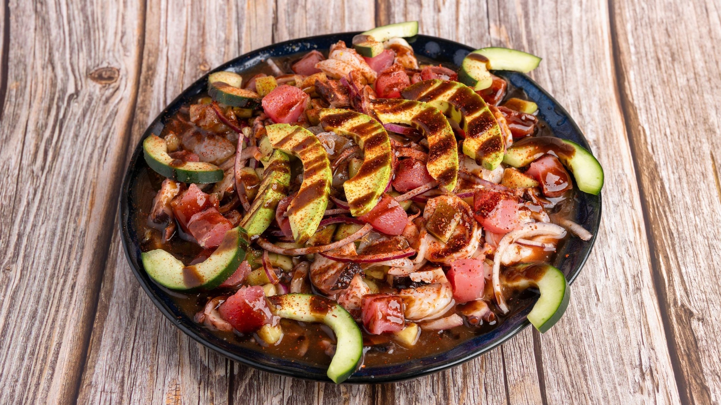 A black plate of mixed ceviche with sliced avocado, tomato, onion, and dark sauce on a wooden table.