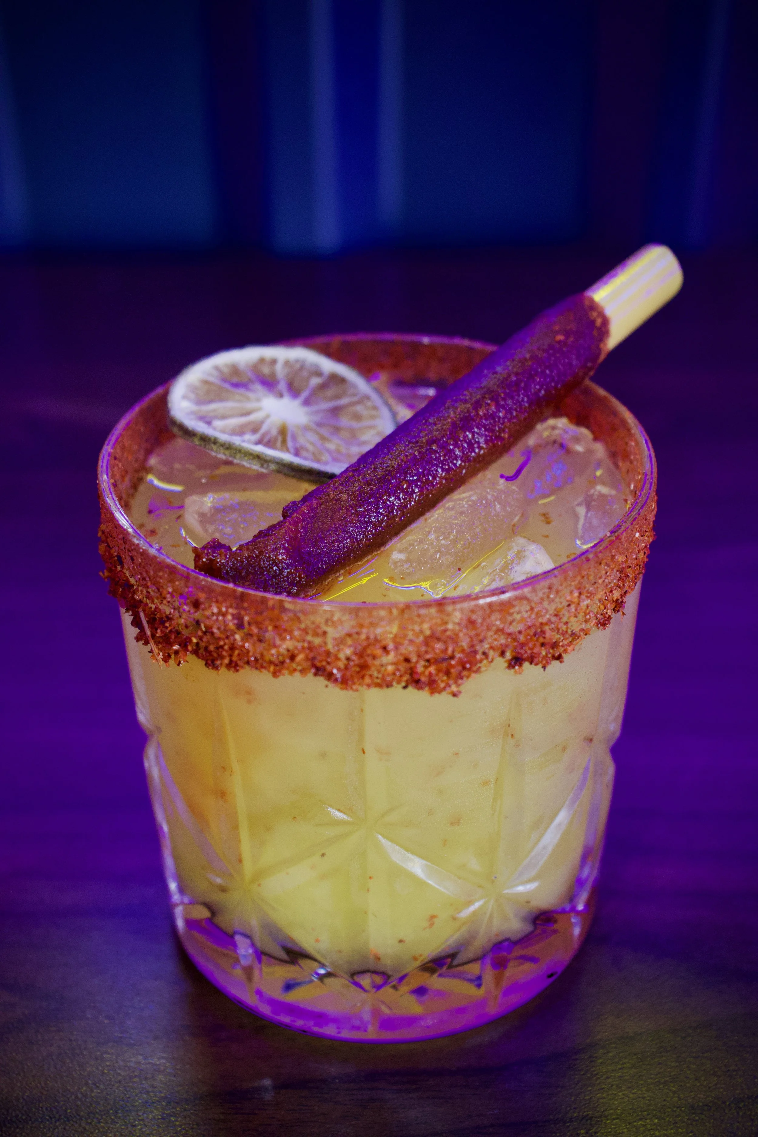 A cocktail drink in a textured glass with ice, garnished with a slice of dried lemon and a cinnamon stick, with a rim coated in chili powder.