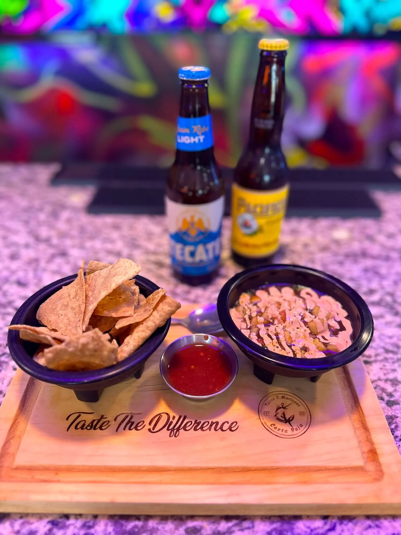 Two small bowls of Mexican food with chips and salsa on a wooden tray, surrounded by two bottles of beer on a colorful, blurry background.