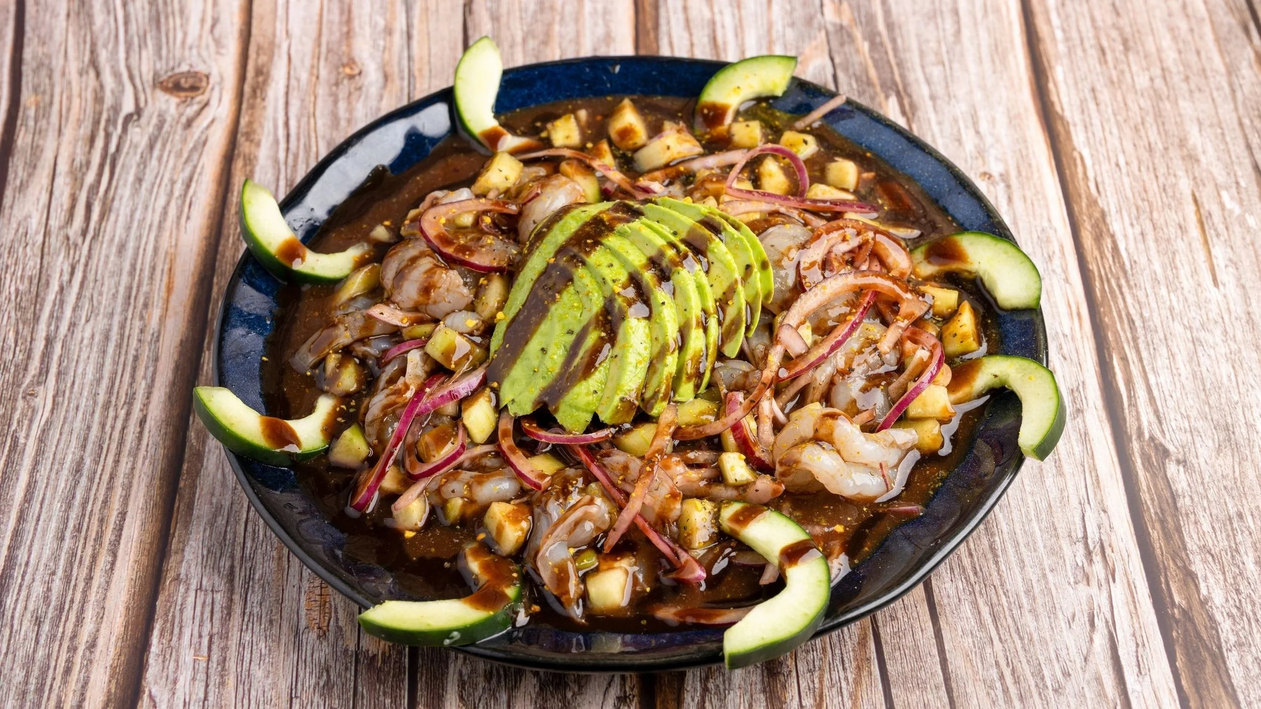 A bowl of shrimp ceviche topped with avocado slices and surrounded by cucumber slices on a wooden table.