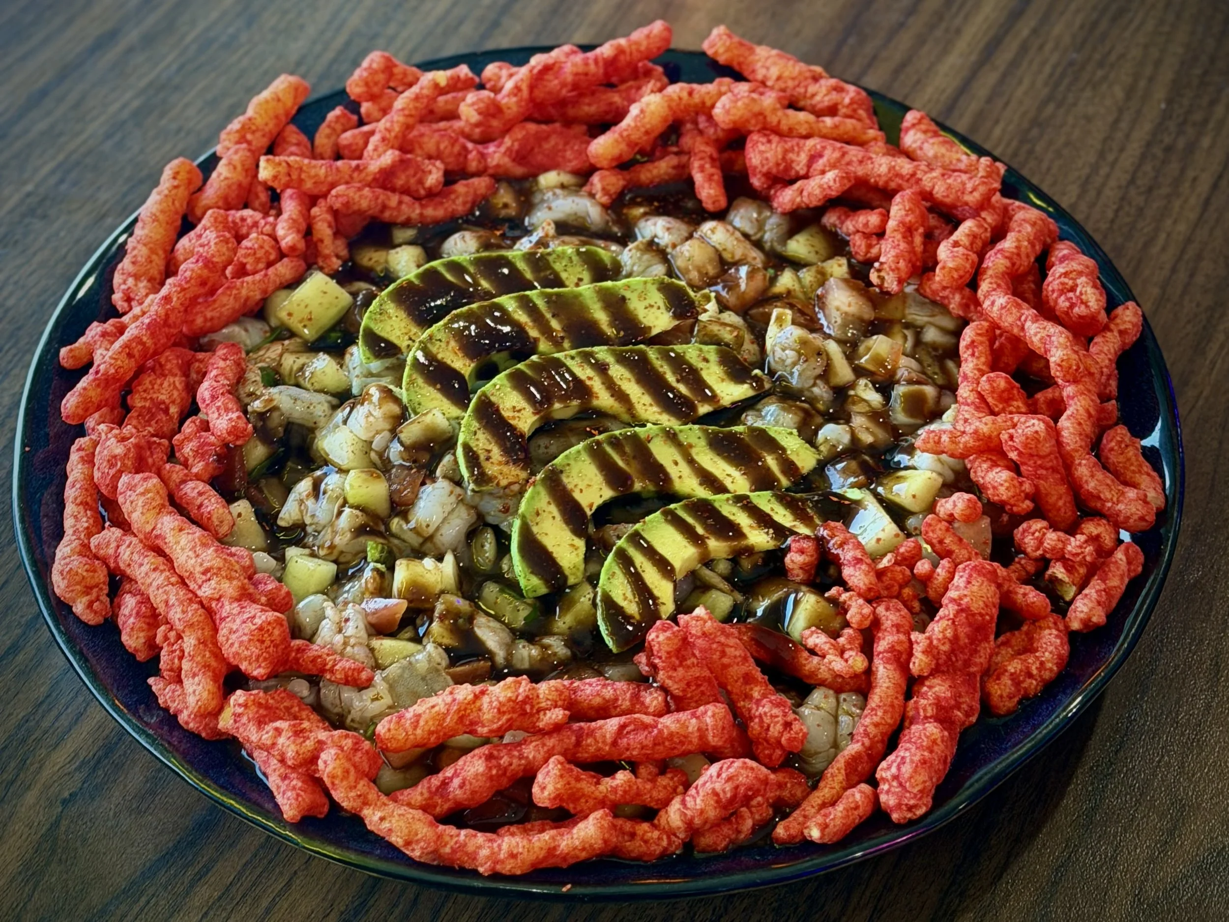 A dish with chopped cucumbers, dark sauce, avocado slices drizzled with sauce, and a red snack border on a black plate.