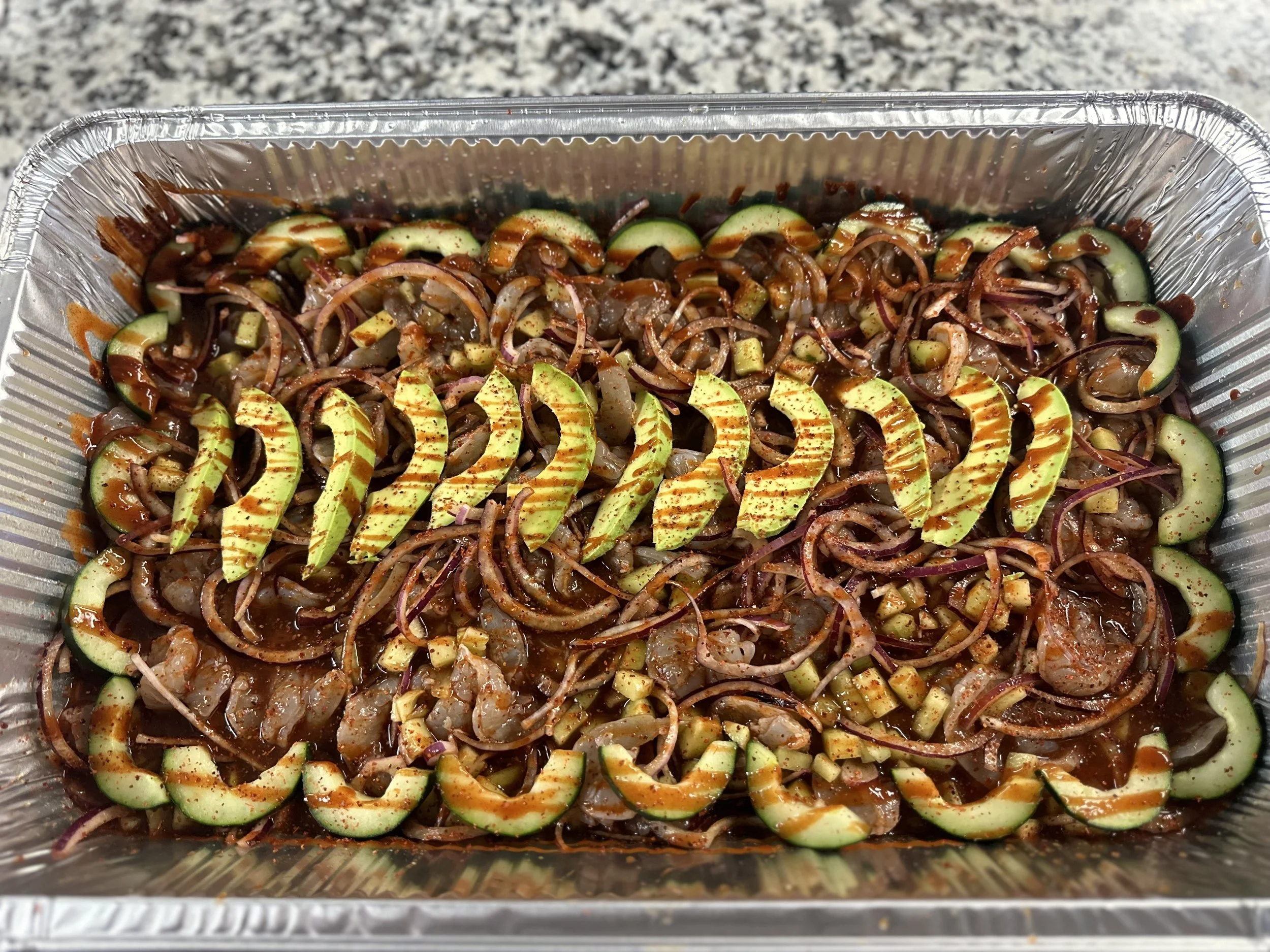A foil container of cooked shrimp with sliced zucchini, onions, and a reddish sauce, possibly hot sauce or seasoning.