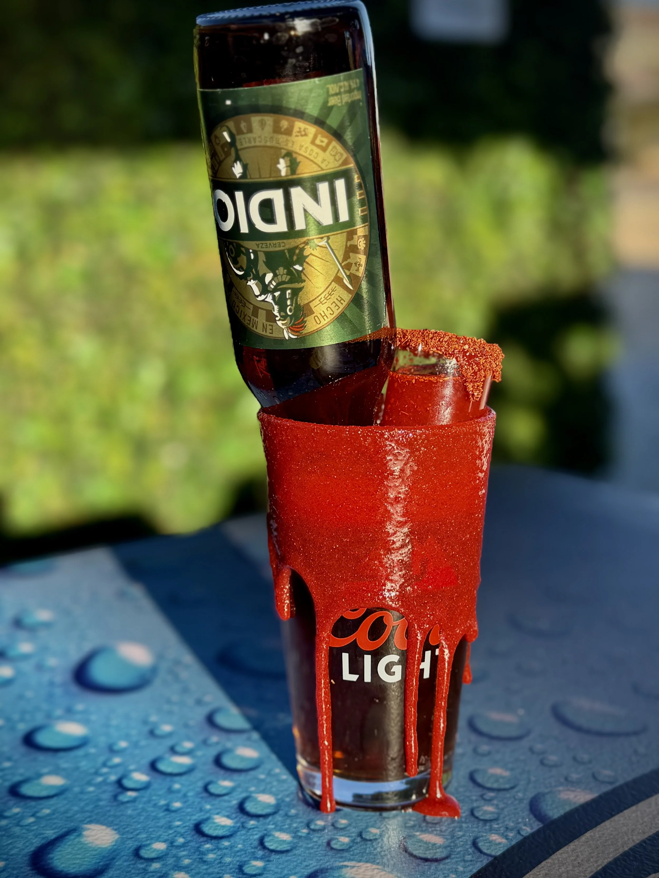 A close-up of a Corona beer bottle with red chili powder on top, set on a surface with water droplets and a blue textured background.