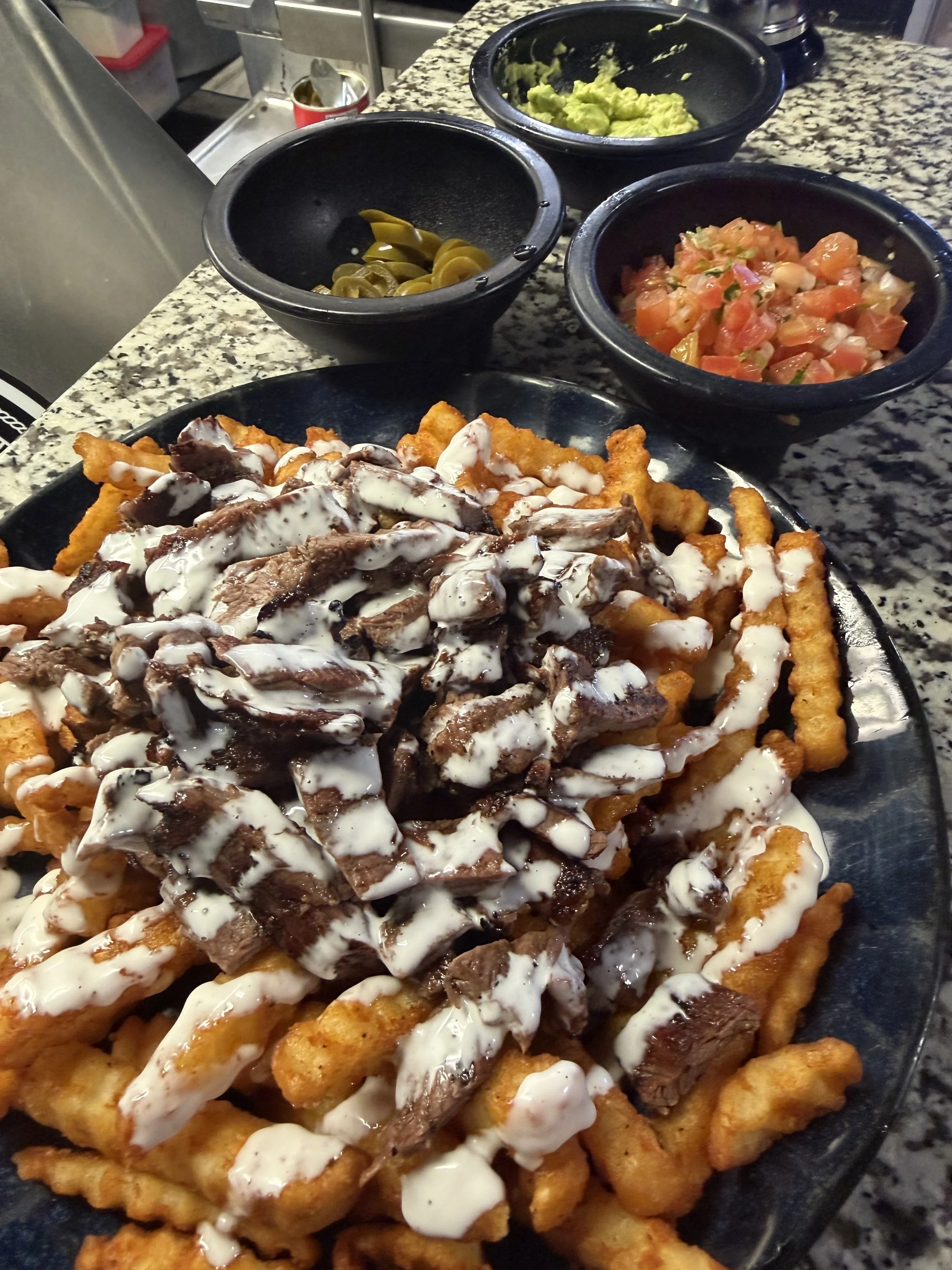 A plate of nachos topped with shredded beef, cheese, and white sauce, with bowls of guacamole, chopped tomatoes, jalapeños, and sliced pickled jalapeños in the background.