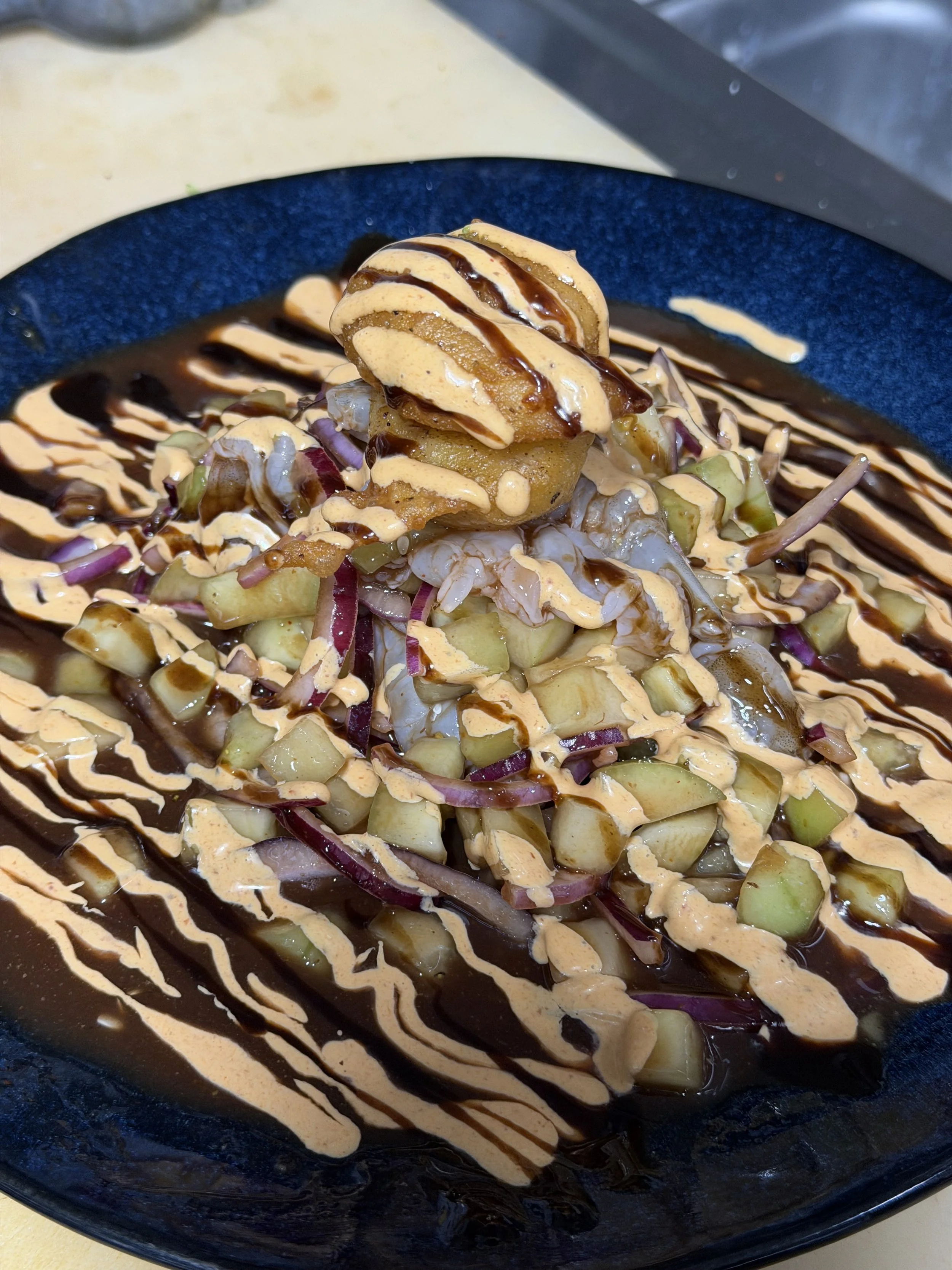 A black bowl holding a salad with chopped cucumbers, red onions, shrimp, and fried plantains topped with drizzles of garlic sauce and a dark sauce, possibly balsamic glaze.