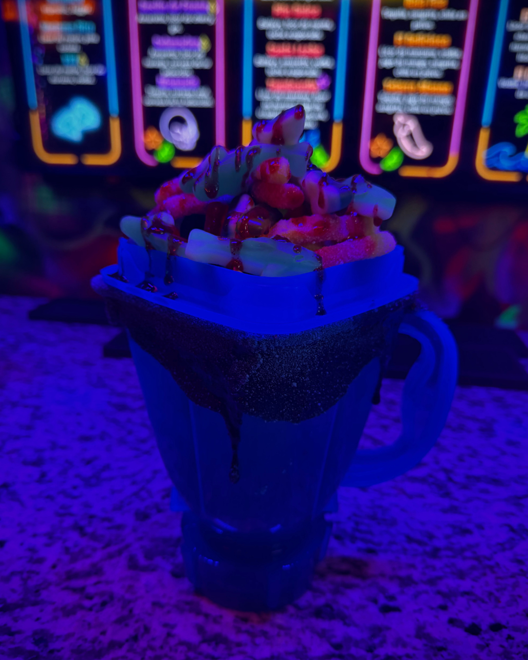 Neon-lit dessert in a mug with whipped cream, strawberry syrup, and toppings on a granite countertop, with a menu in the background.