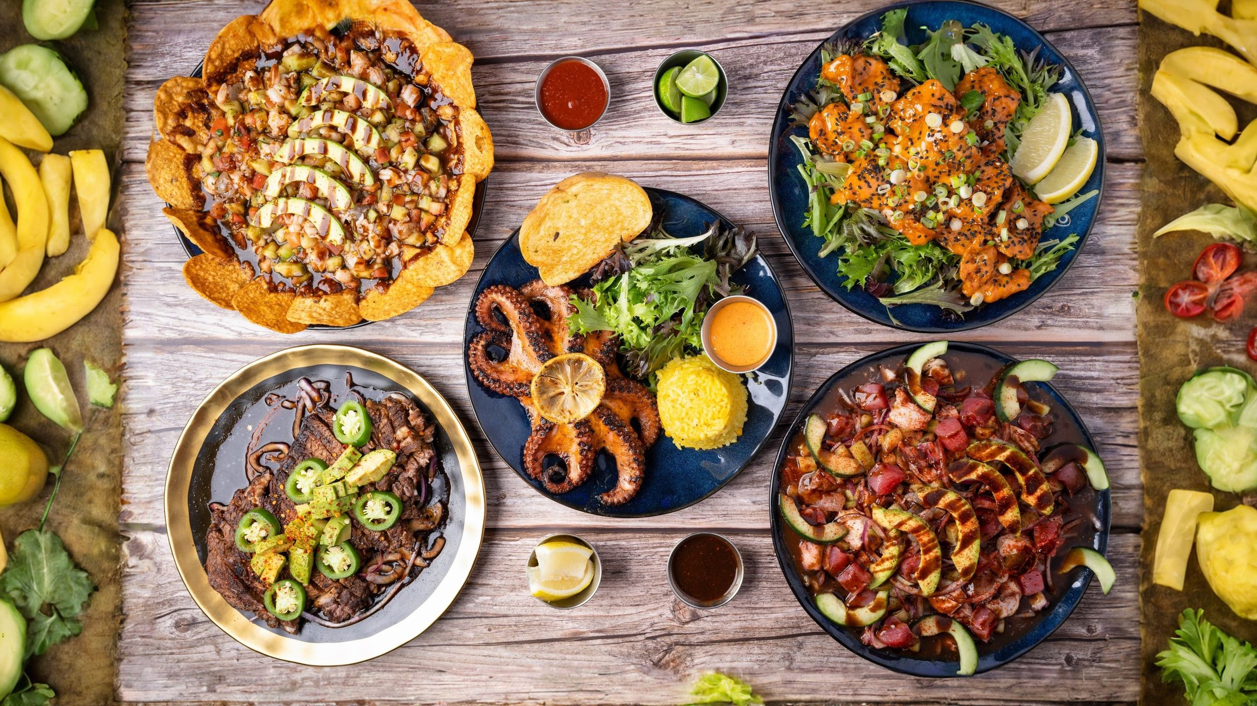 A variety of Mexican dishes including tacos, salad with salmon, and grilled meat, surrounded by fresh vegetables and condiments on a wooden table.