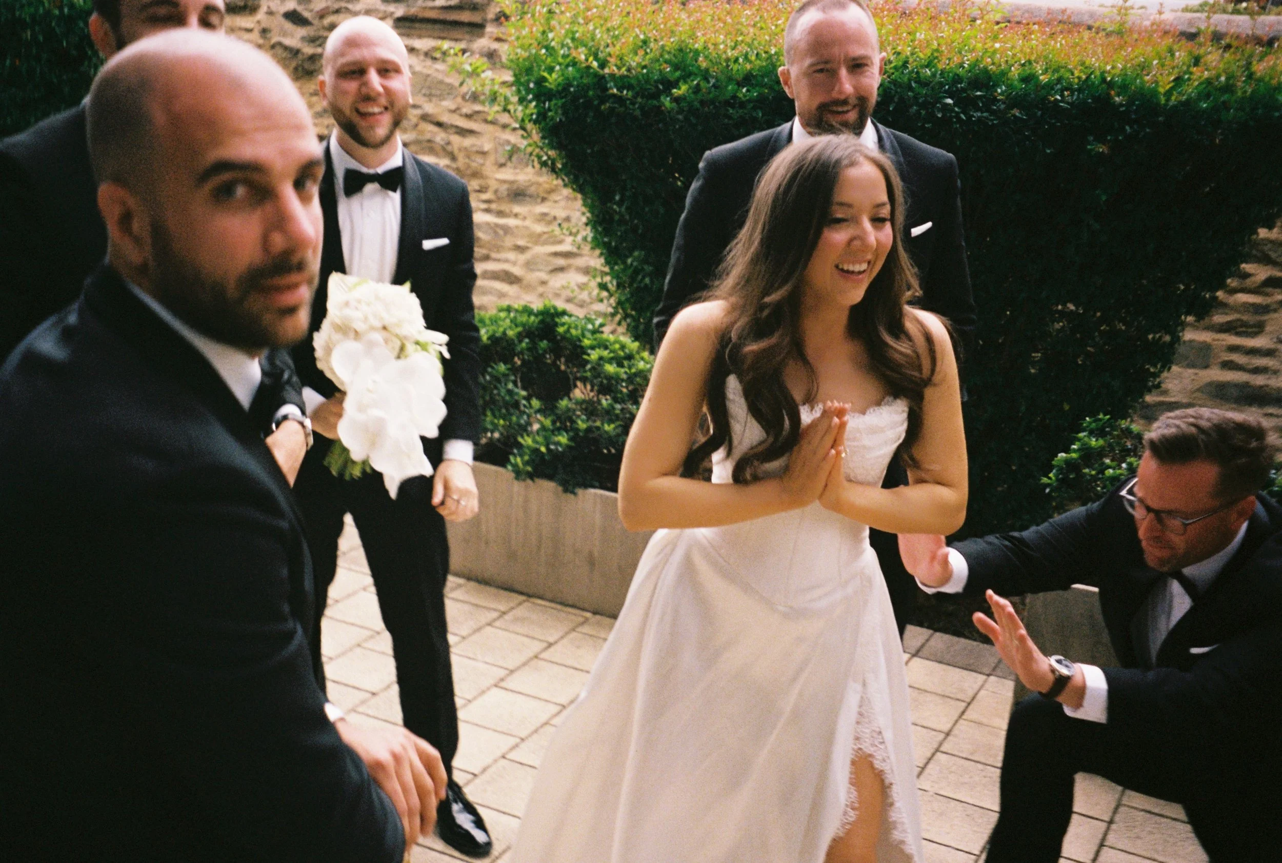 A bride in a white wedding dress smiling and covering her hands together, surrounded by five men in suits, some smiling and one holding a bouquet of white flowers, outdoors with stone and greenery background.