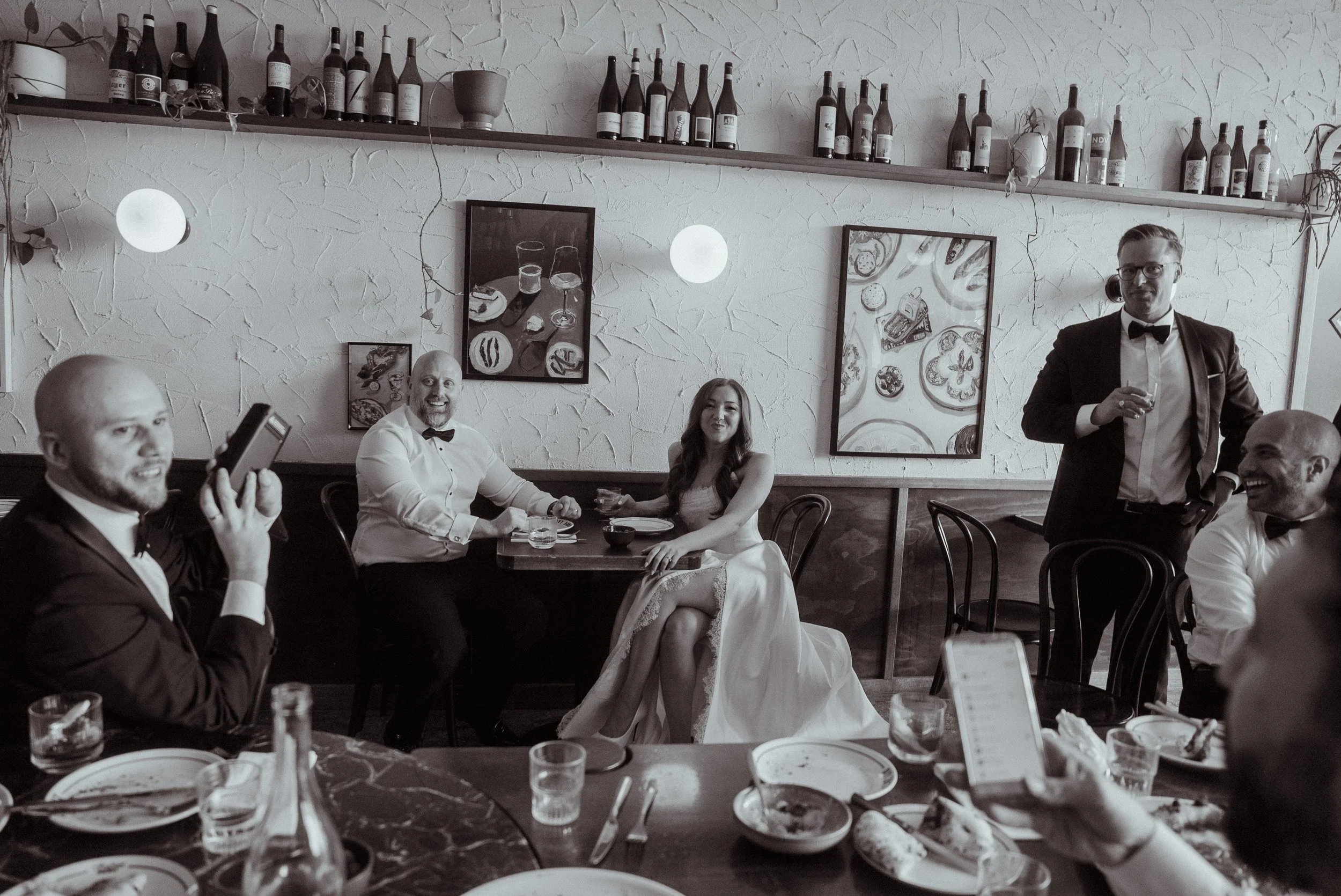 A black-and-white photo of a wedding celebration in a restaurant, featuring a woman in a white wedding dress sitting at a table with friends and family, some of whom are wearing tuxedos, with bottles on a shelf above and framed artwork on the wall.