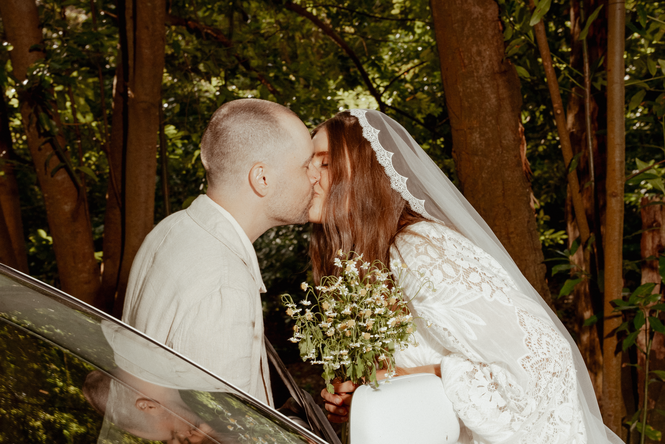 A bride and groom kiss outdoors next to a car, with trees in the background. The bride is holding a bouquet of small white flowers and wearing a lace wedding dress with a veil.