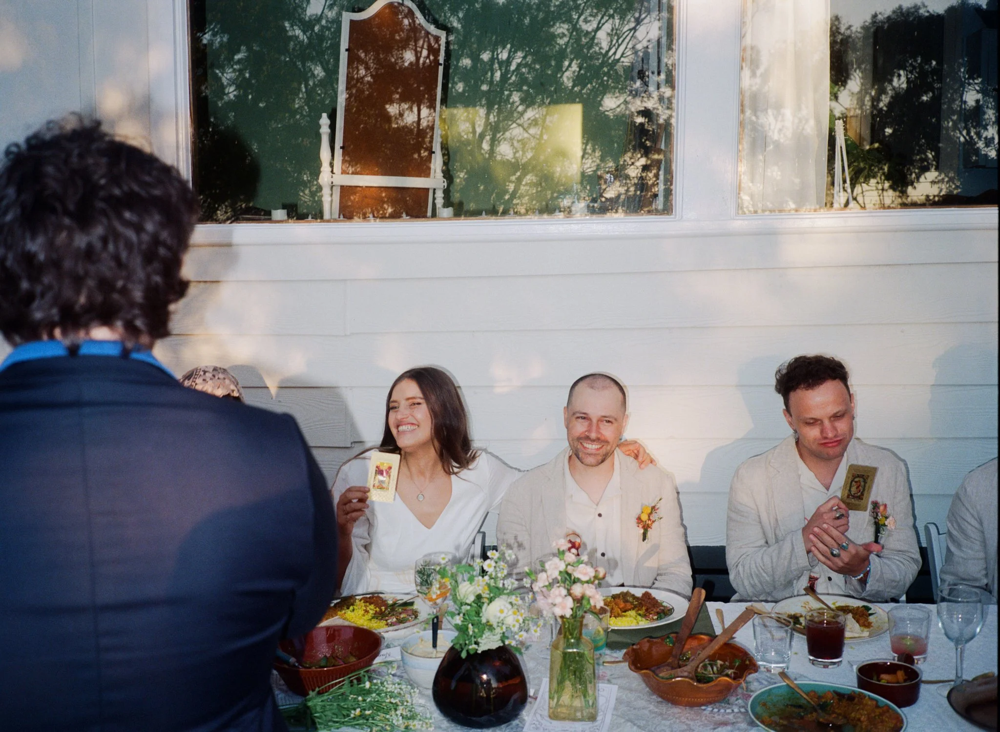 Group of people at a dinner table, celebrating with food and flowers, two people holding tarot cards, smiling and enjoying the gathering.