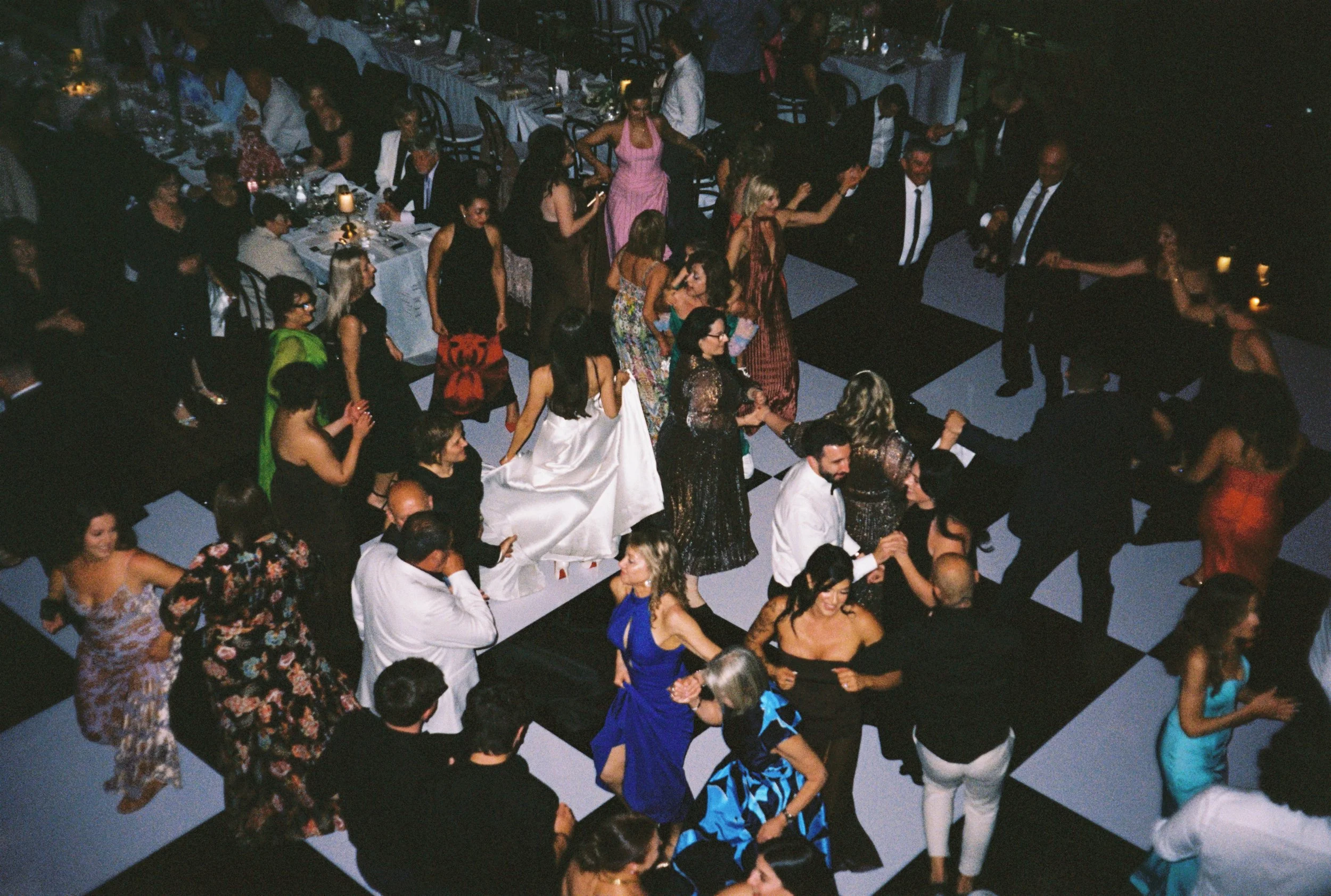 People dancing on a checkered dance floor at a formal event with round tables and seated guests in the background.
