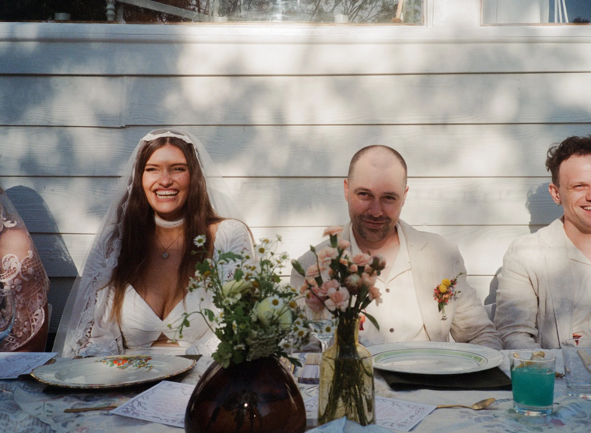 People sitting at a decorated table outdoors, including a smiling woman with long hair wearing a wedding dress and veil, and two men in light-colored suits, with flowers and plates on the table.