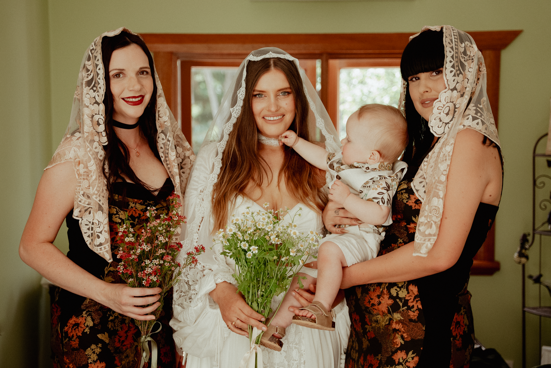 Four women, one holding a baby, are dressed in floral and lace clothing with lace headscarves. The woman in the center is wearing a white dress and veil, smiling and holding a bouquet of green flowers. The other women are holding smaller flower bouqu