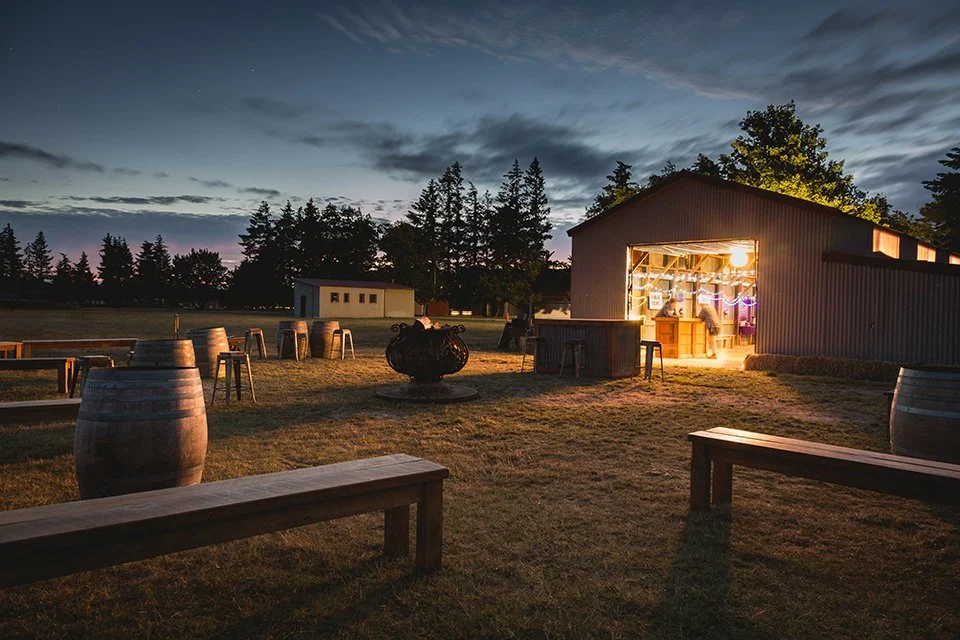 A rustic outdoor gathering area at sunset with a barn illuminated inside, surrounded by seating and barrels, and a fire pit in the center, with trees and a partly cloudy sky in the background.