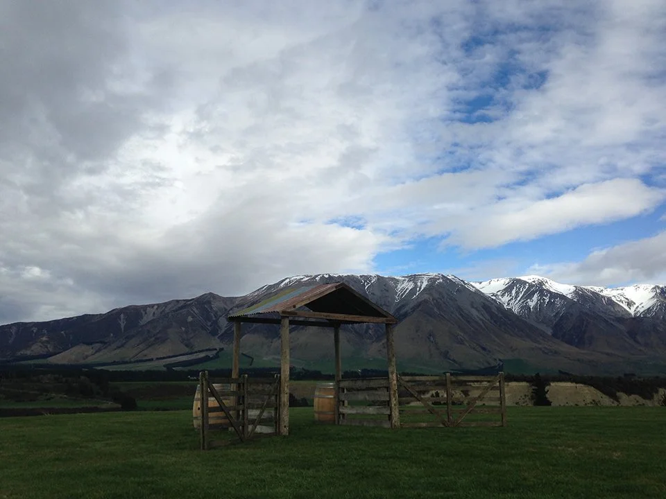 A rural landscape with a wooden shelter, green grass, and a mountain range with snow-capped peaks under a partly cloudy sky.