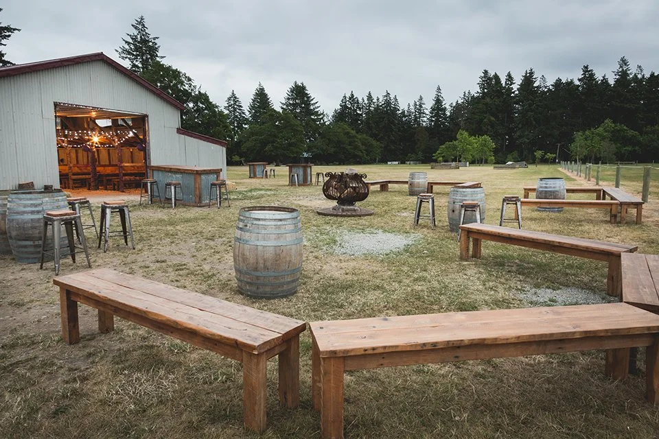 Outdoor patio area with wooden benches, barrels, and tables in front of a barn, with trees and grass in the background, under cloudy sky.